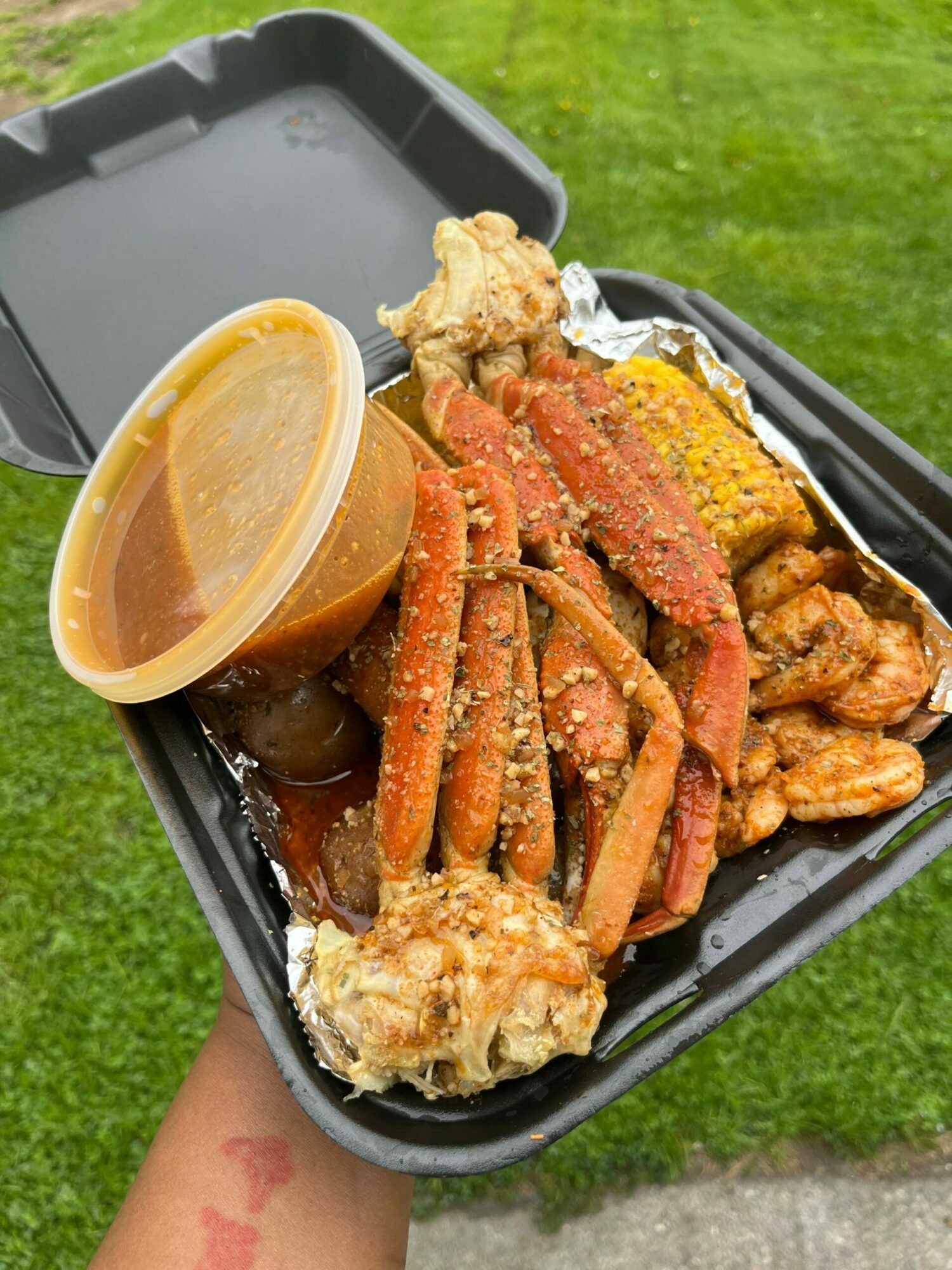 Tray of cooked seafood, corn, and sauce on grass, held by a person with a tattooed arm.
