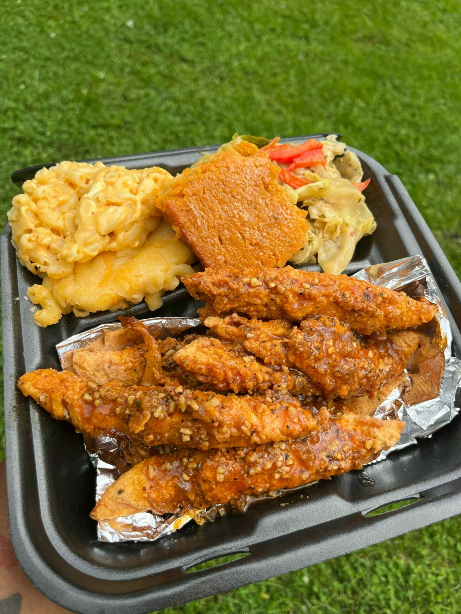 Fried fish fillets, fried chicken, mashed potatoes, and vegetables on a black tray outdoors.