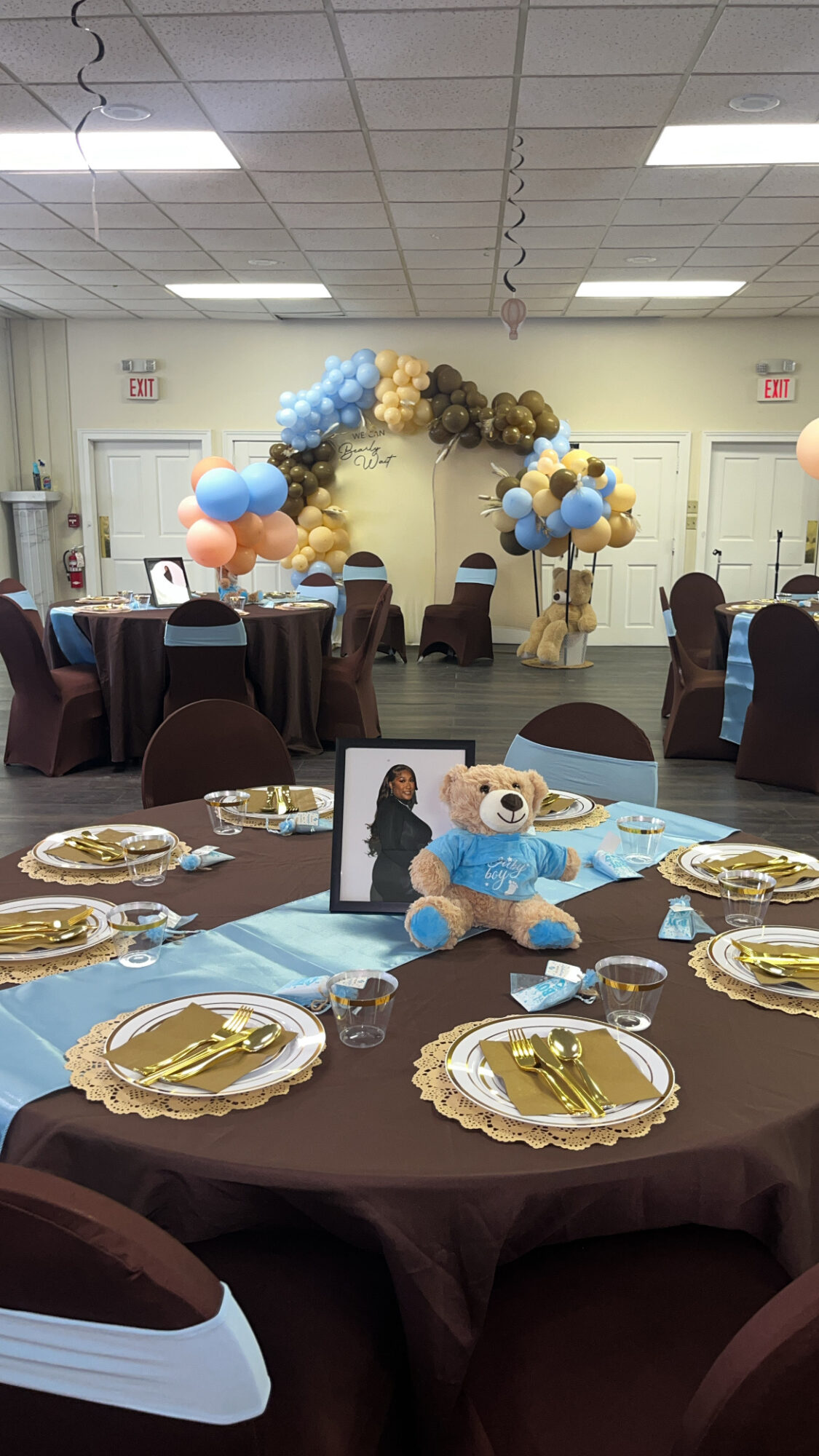 Party room with decorated balloon arch, tables set with gold plates, and a teddy bear centerpiece, ceiling lights overhead.