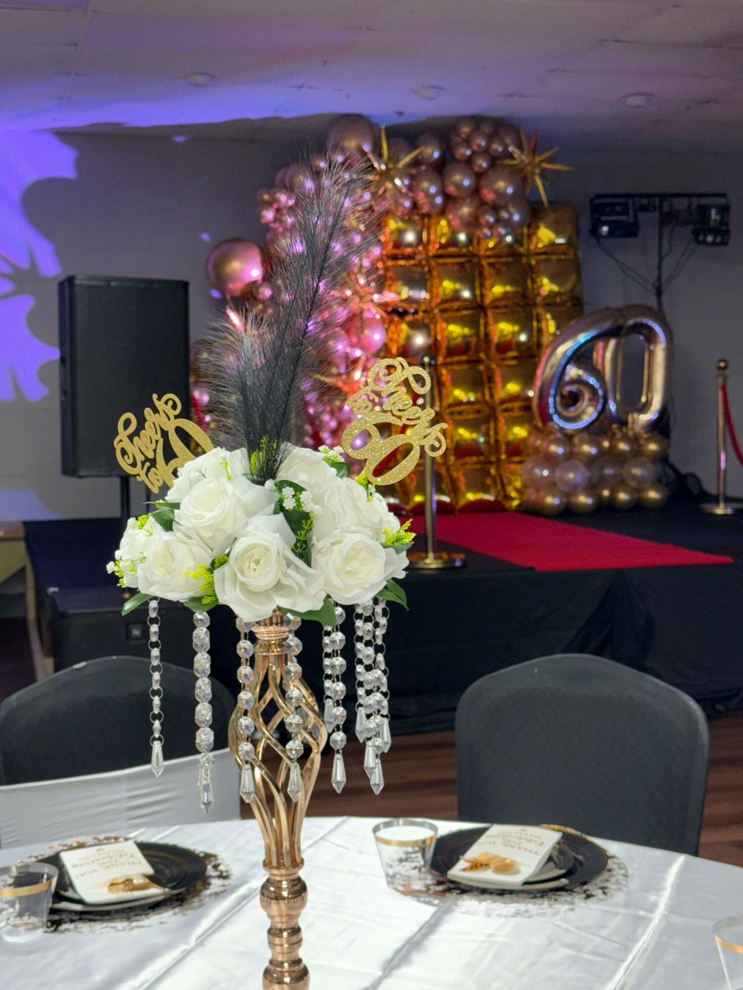 Table with white flowers, gold decorations, and a dessert display in the background, with a black chair and tableware visible.