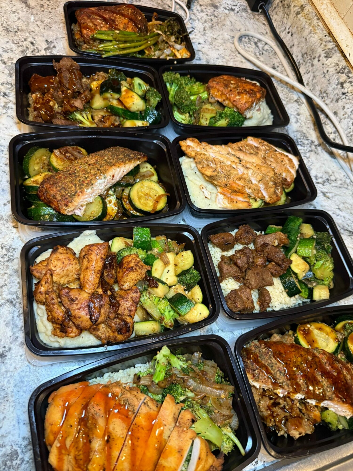 Multiple black meal containers filled with various cooked foods, including grilled meats, vegetables, and seafood, on a countertop.