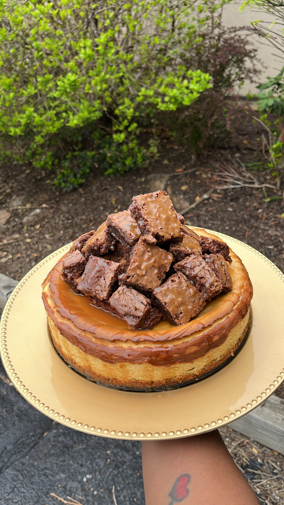 A dessert with a glazed base topped with chocolate chunks on a gold plate, outdoors with greenery in the background.
