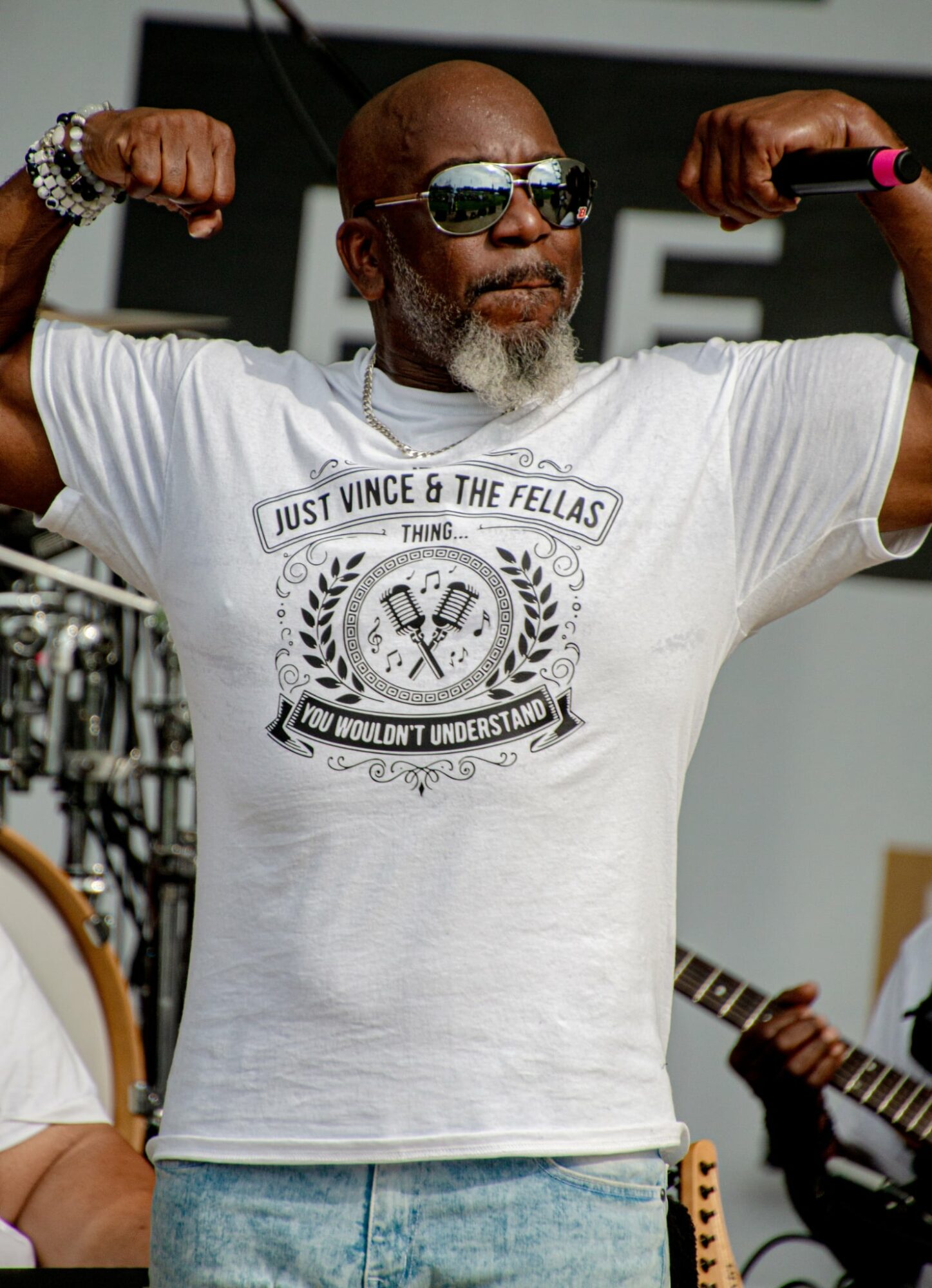 Man with sunglasses flexing arms, wearing a white T-shirt with a graphic design, in an outdoor setting.