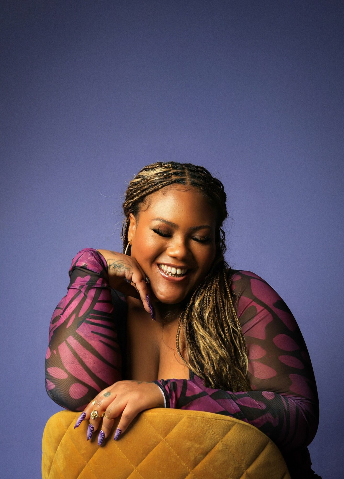 Smiling woman with braided hair, wearing a purple patterned top, resting her chin on her hand, leaning on a yellow cushion.
