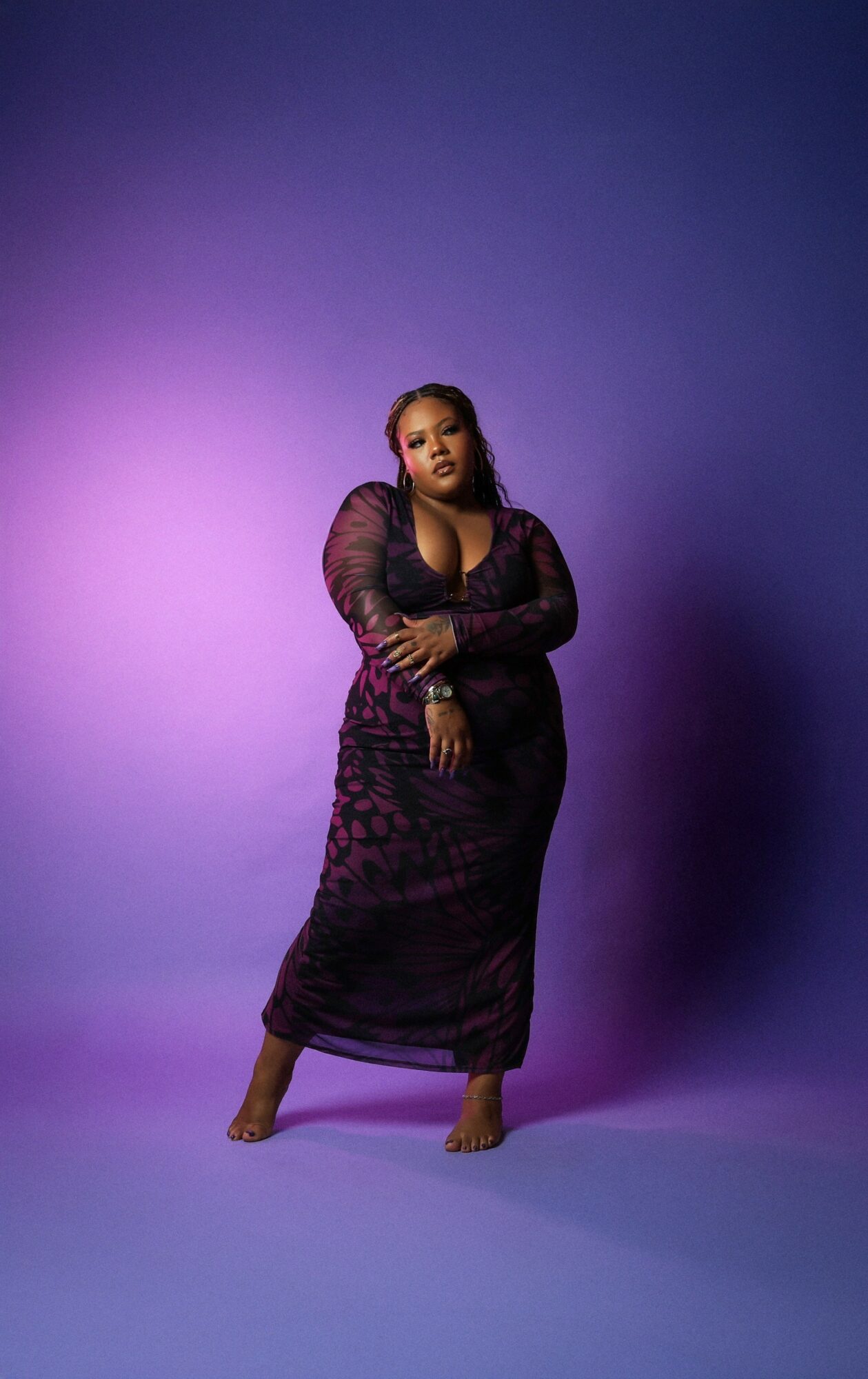 Woman in a long, dark dress standing with arms crossed against a purple gradient background.
