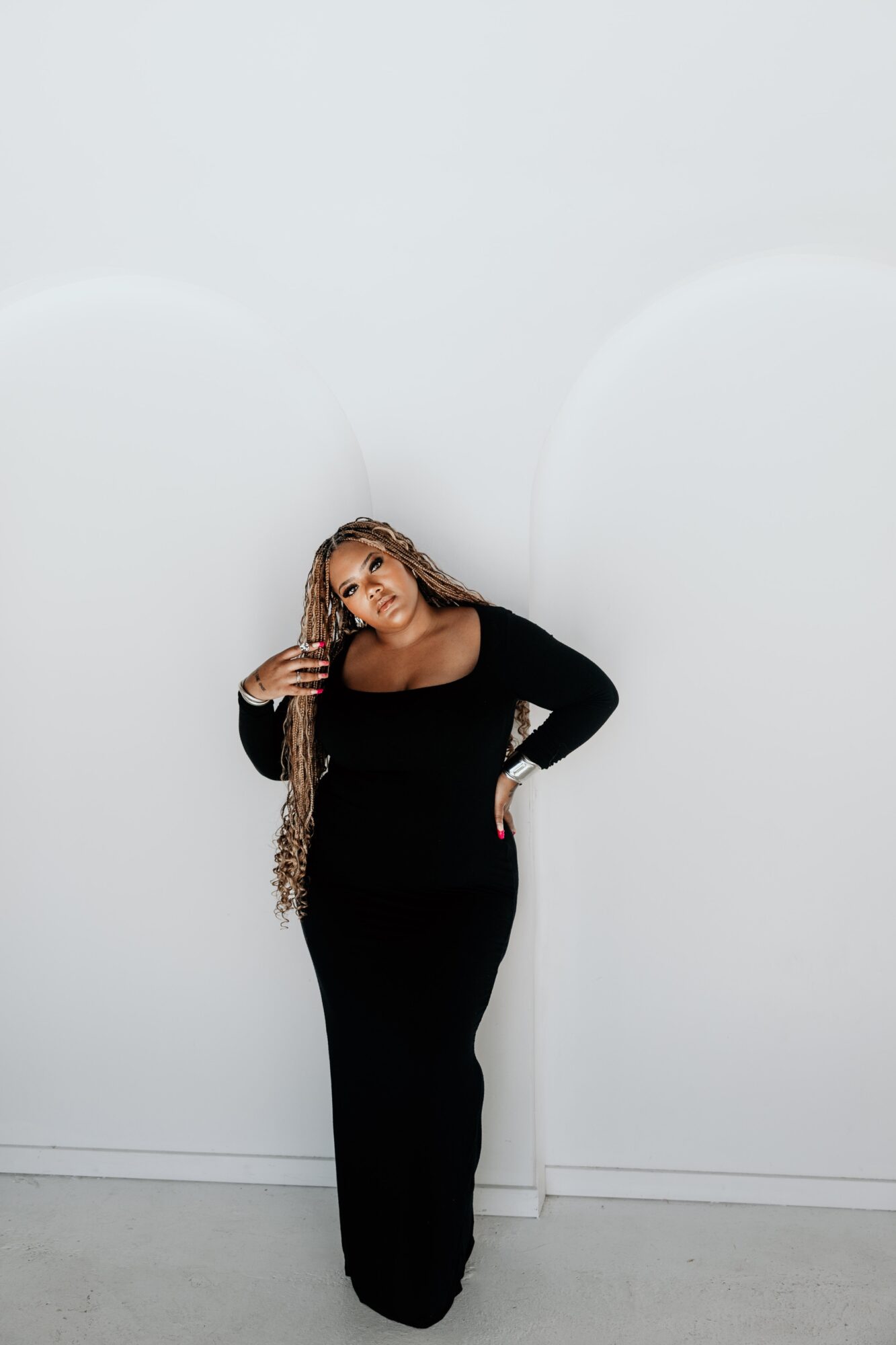 Woman in black dress standing against a plain white wall, with one hand on her hip and the other touching her hair.