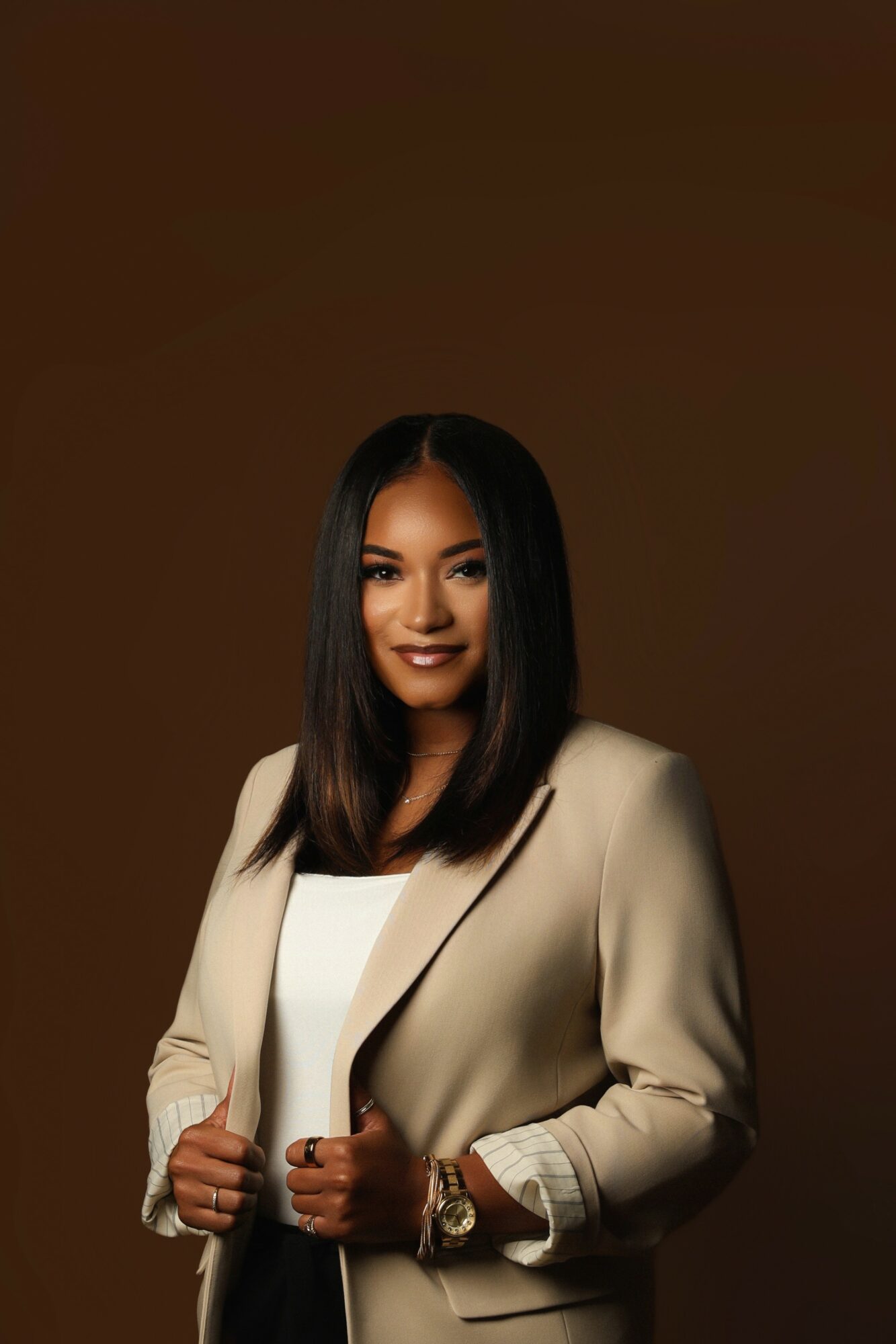 Woman with shoulder-length dark hair wearing a beige blazer and white top, smiling, against a brown background.
