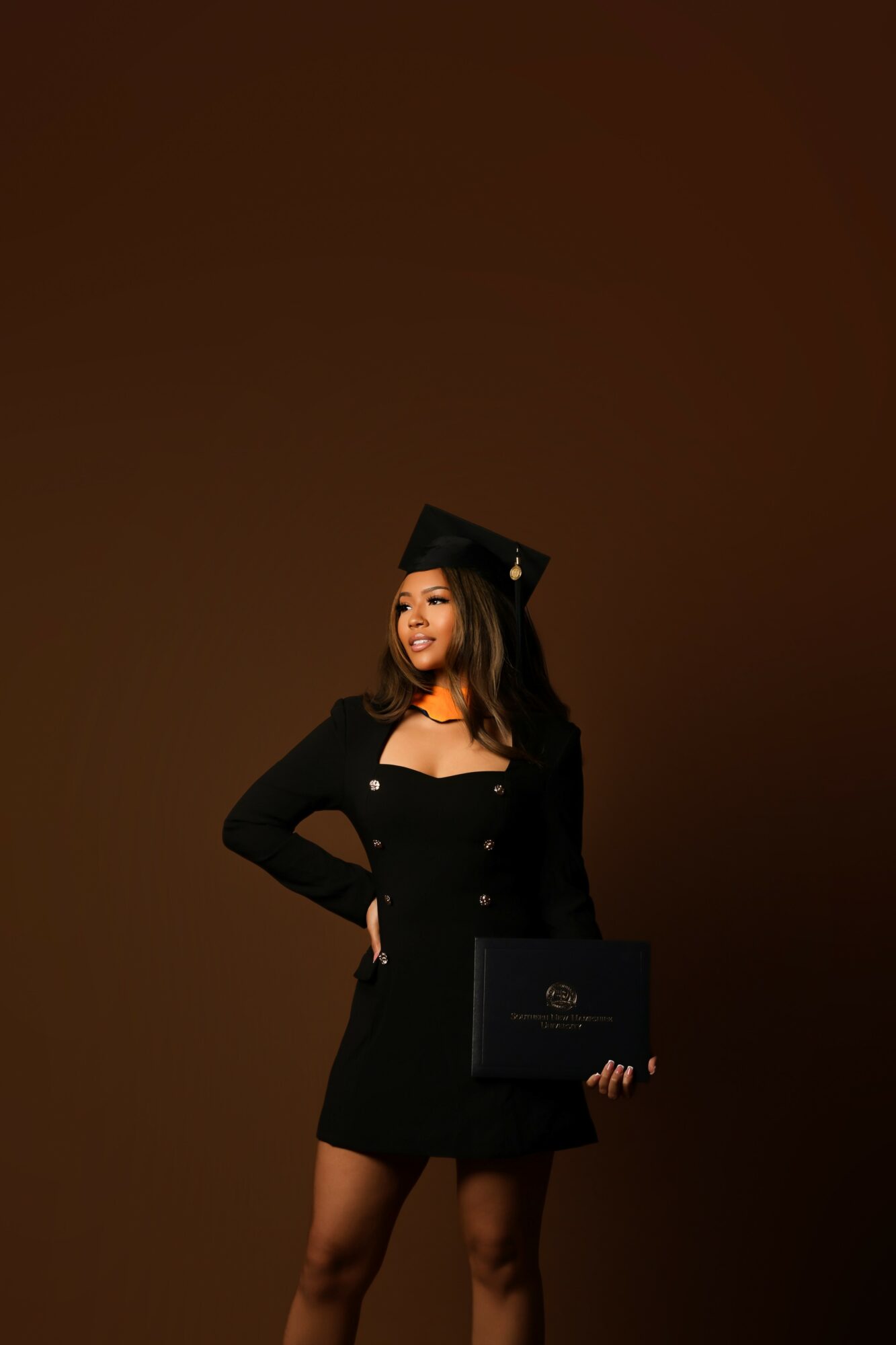Young woman in graduation cap and gown holding diploma, smiling, against a plain brown background.