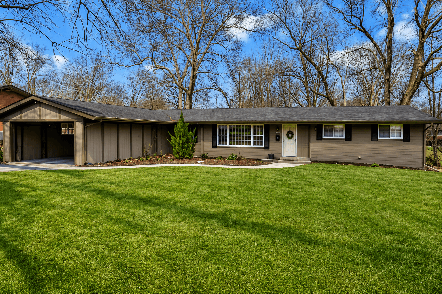 Single-story house with a brown exterior, white window frames, and a front door, surrounded by a green lawn and trees.