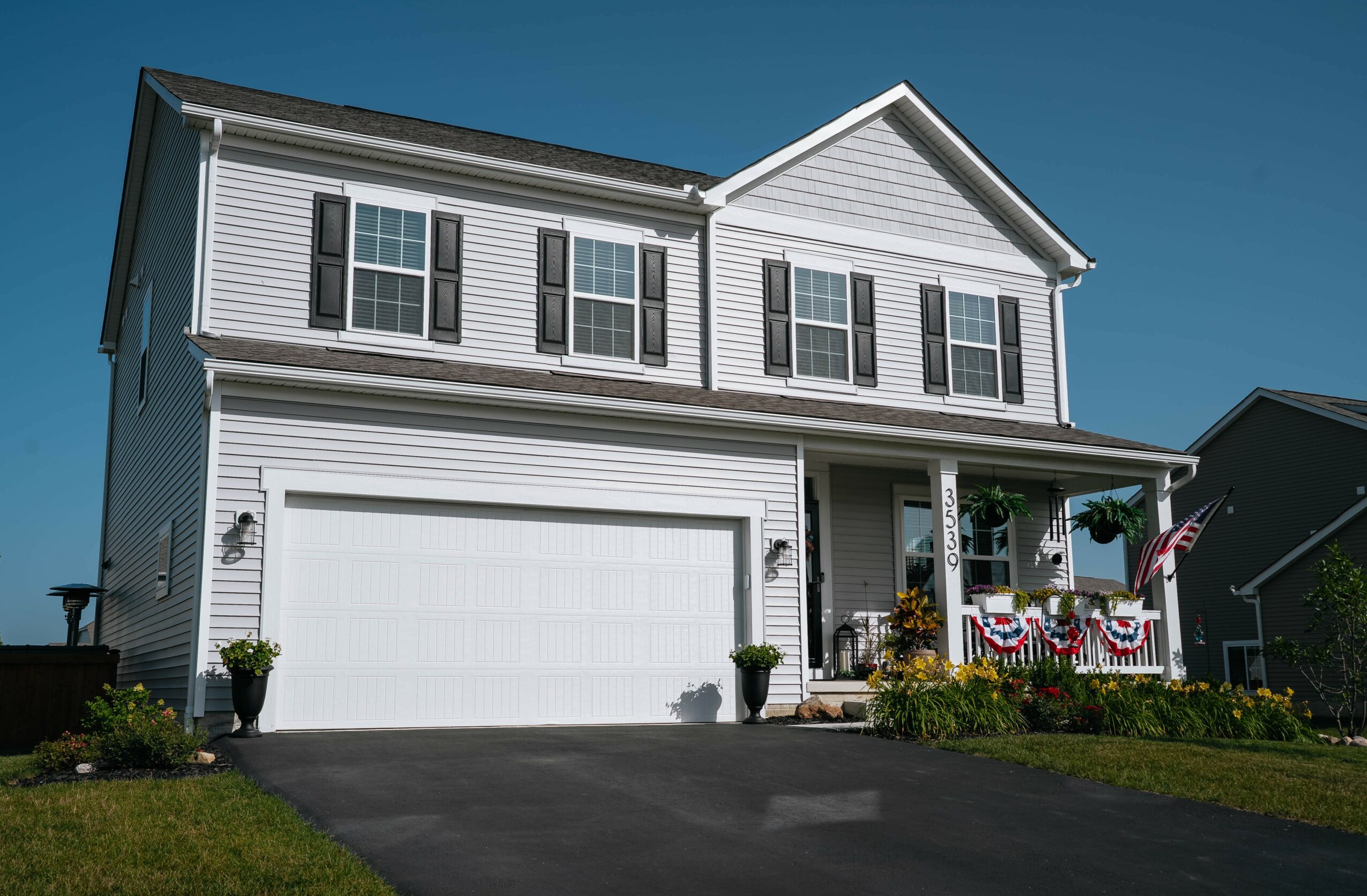 Two-story house with white siding, black shutters, a front porch with chairs, and a two-car garage, under a clear blue sky.