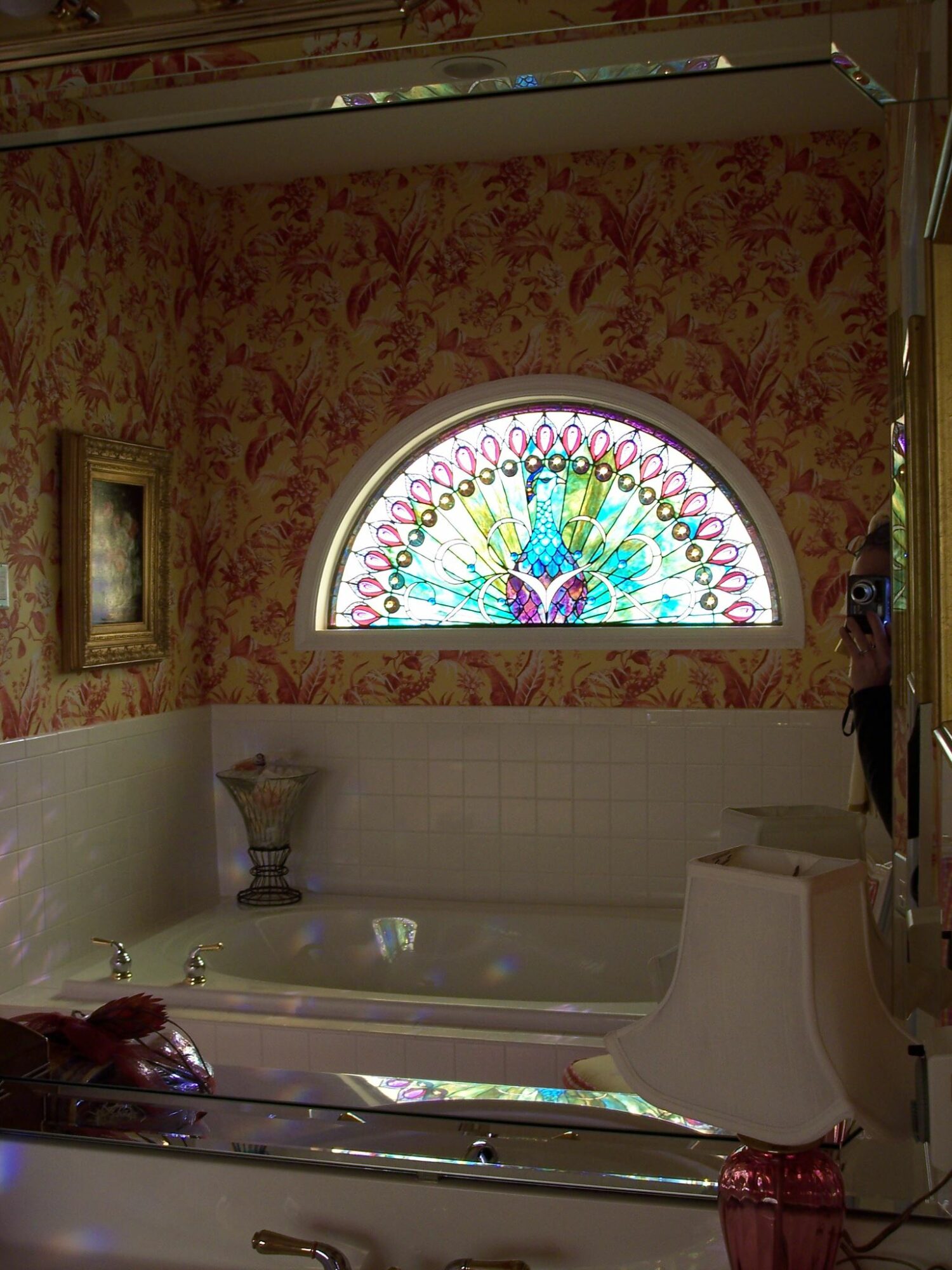 Bathroom with a stained glass window of a peacock, floral wallpaper, and a bathtub with a faucet and a toilet.