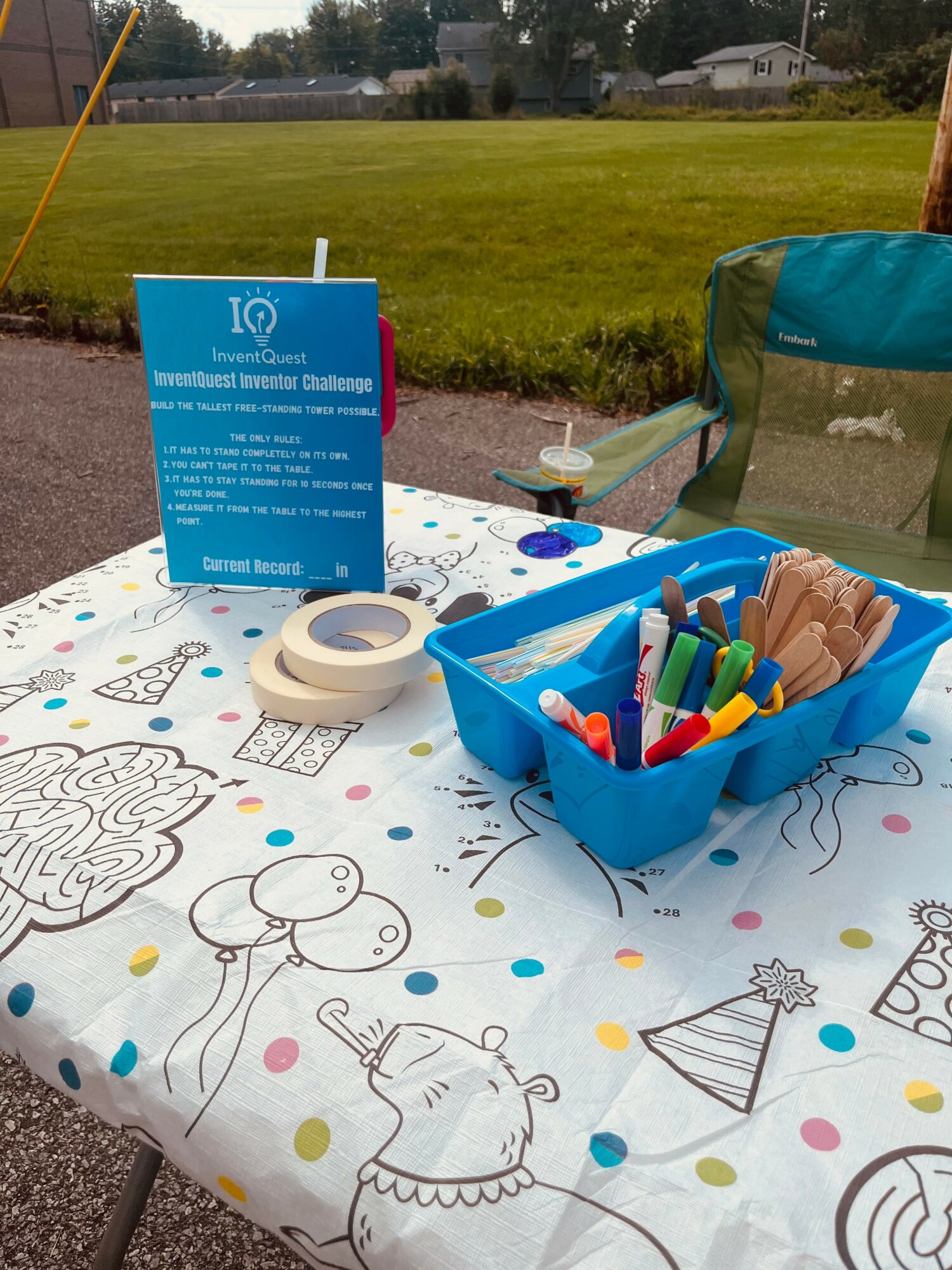 Table with markers, a blue container, and a sign outdoors, with grass and trees in the background.