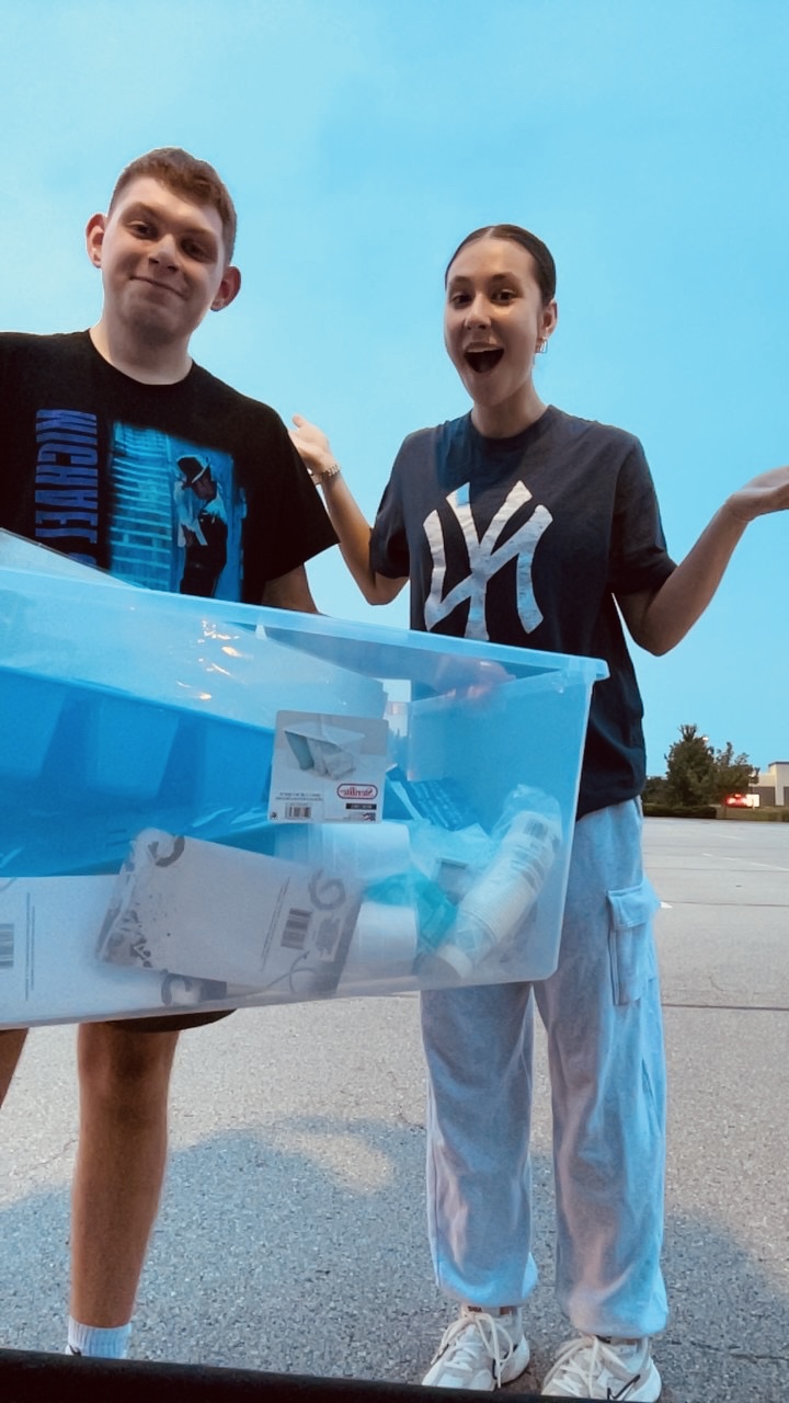 Two people standing outdoors, one holding a large transparent container filled with paper, sky in background.