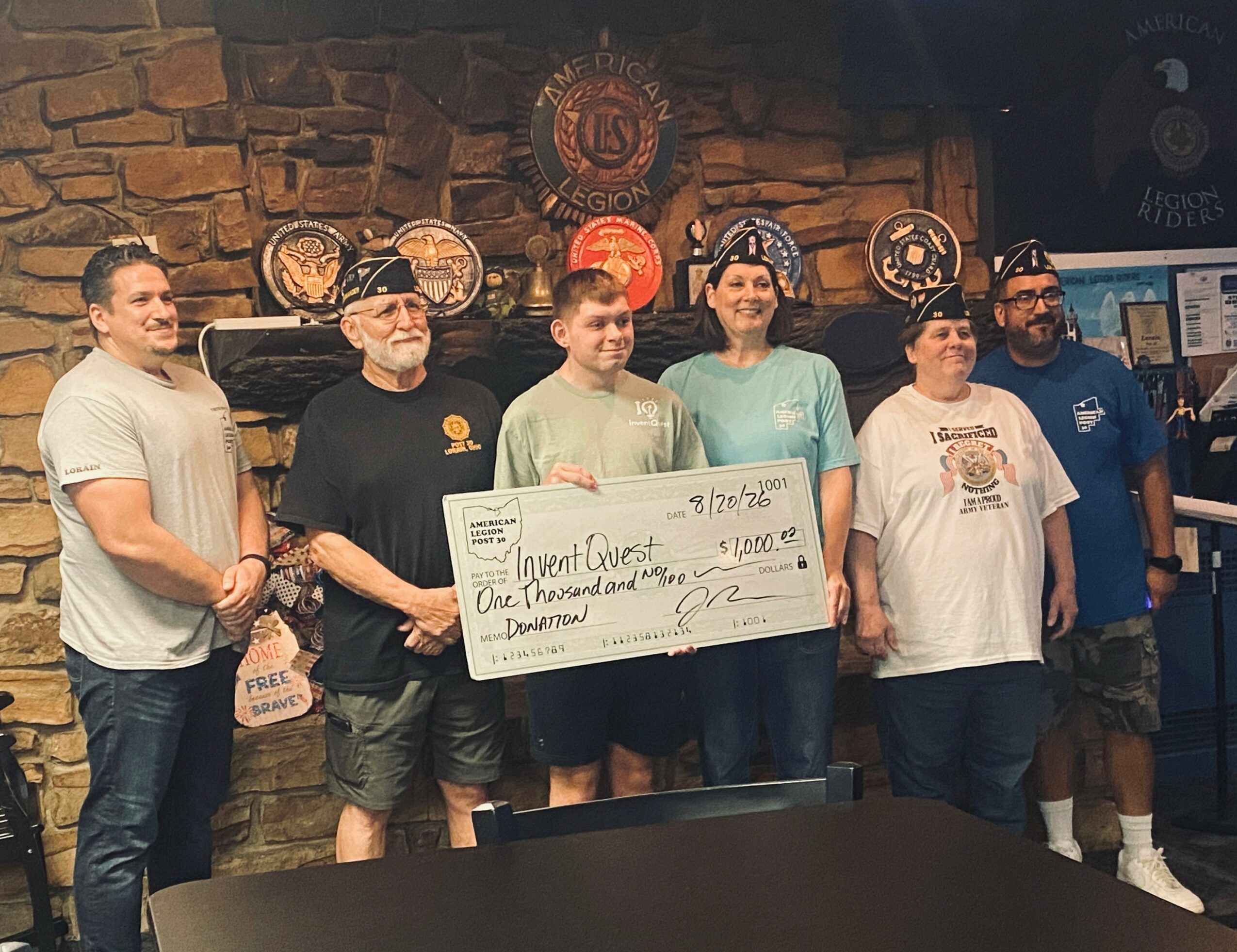 Group of six people standing indoors, holding a large check, with a brick wall and decorative plates behind them.