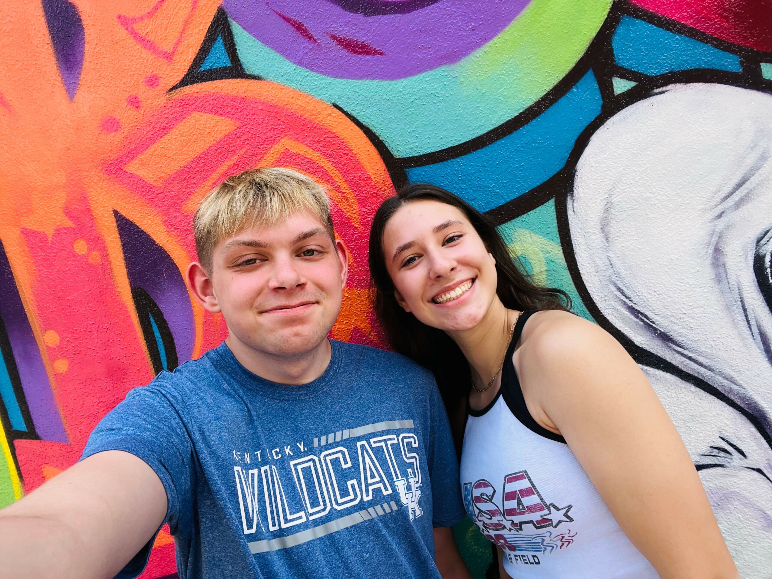 Two smiling people taking a selfie in front of a colorful graffiti wall.