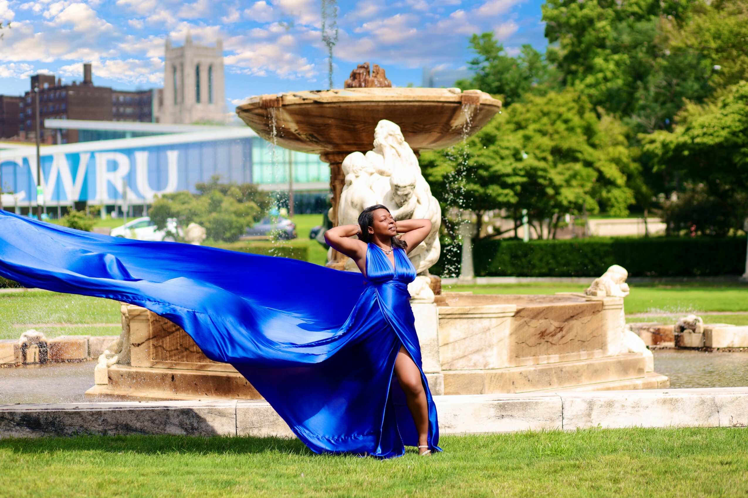 Person in a flowing blue dress standing in front of a fountain with a statue, trees, and buildings in the background.