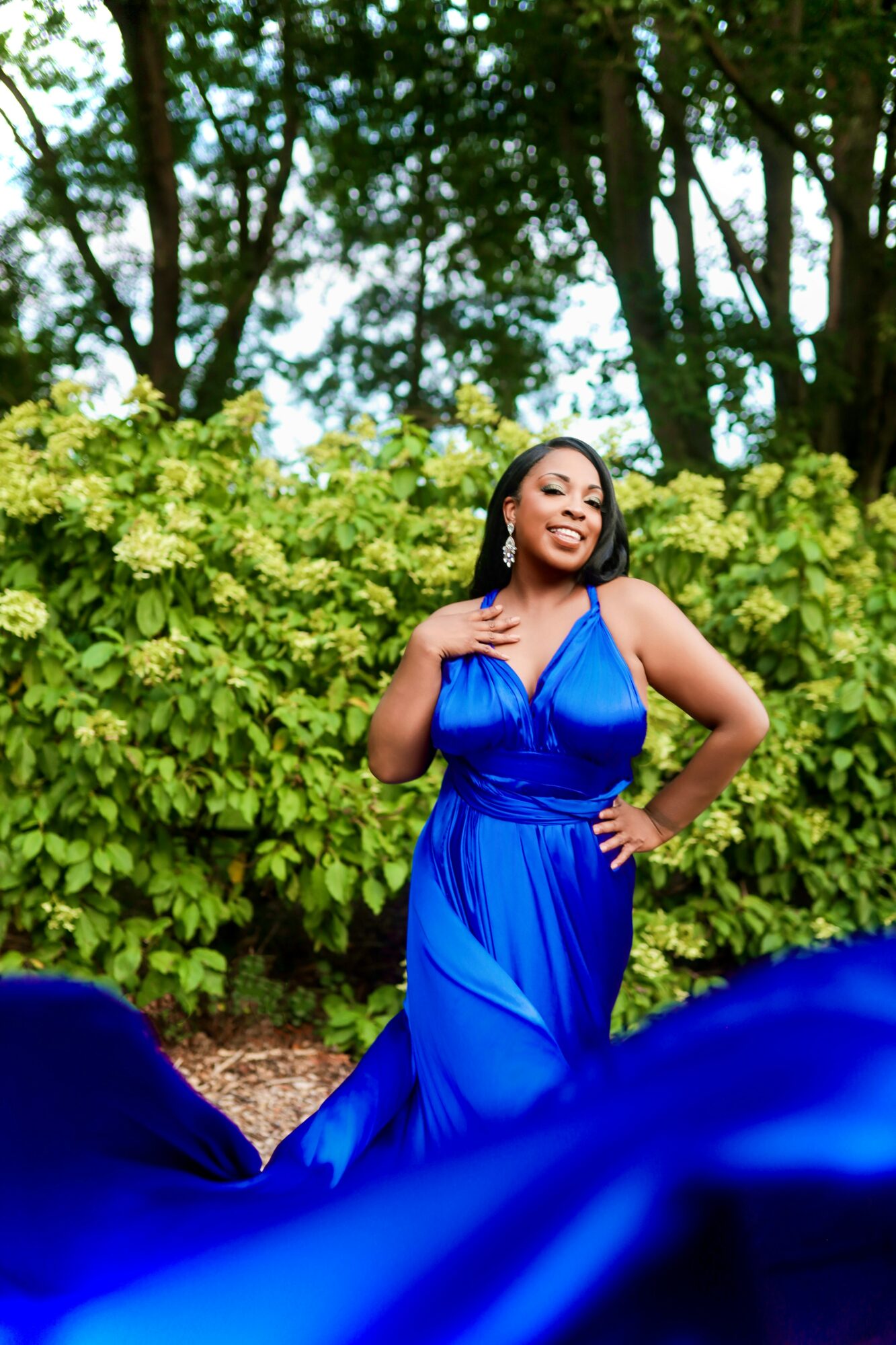 Woman in a blue gown standing outdoors with green foliage background, smiling, hand on waist, flowing fabric at bottom.