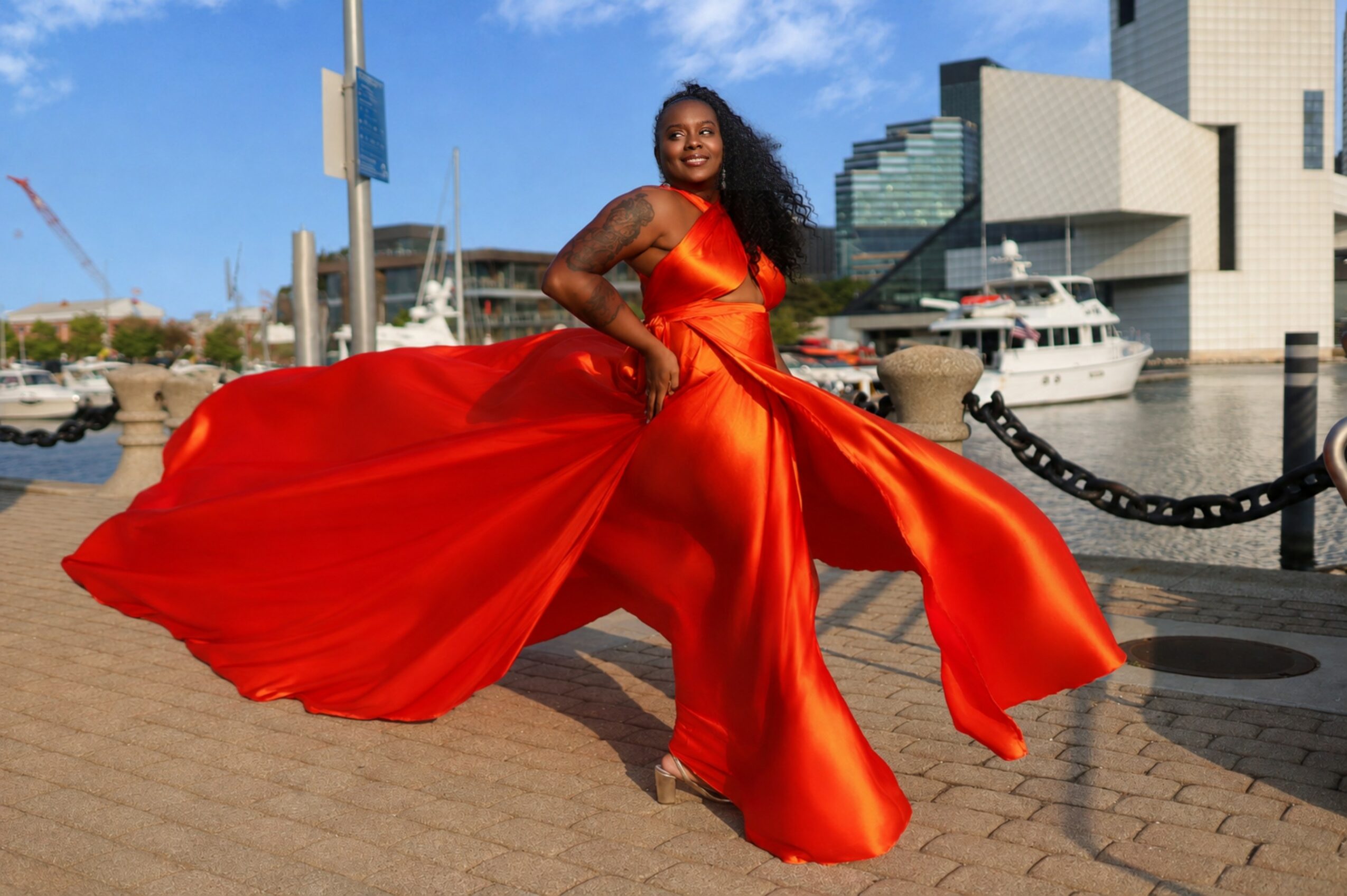 Woman in a flowing red dress dancing outdoors near water and modern buildings, smiling, with a blue sky.