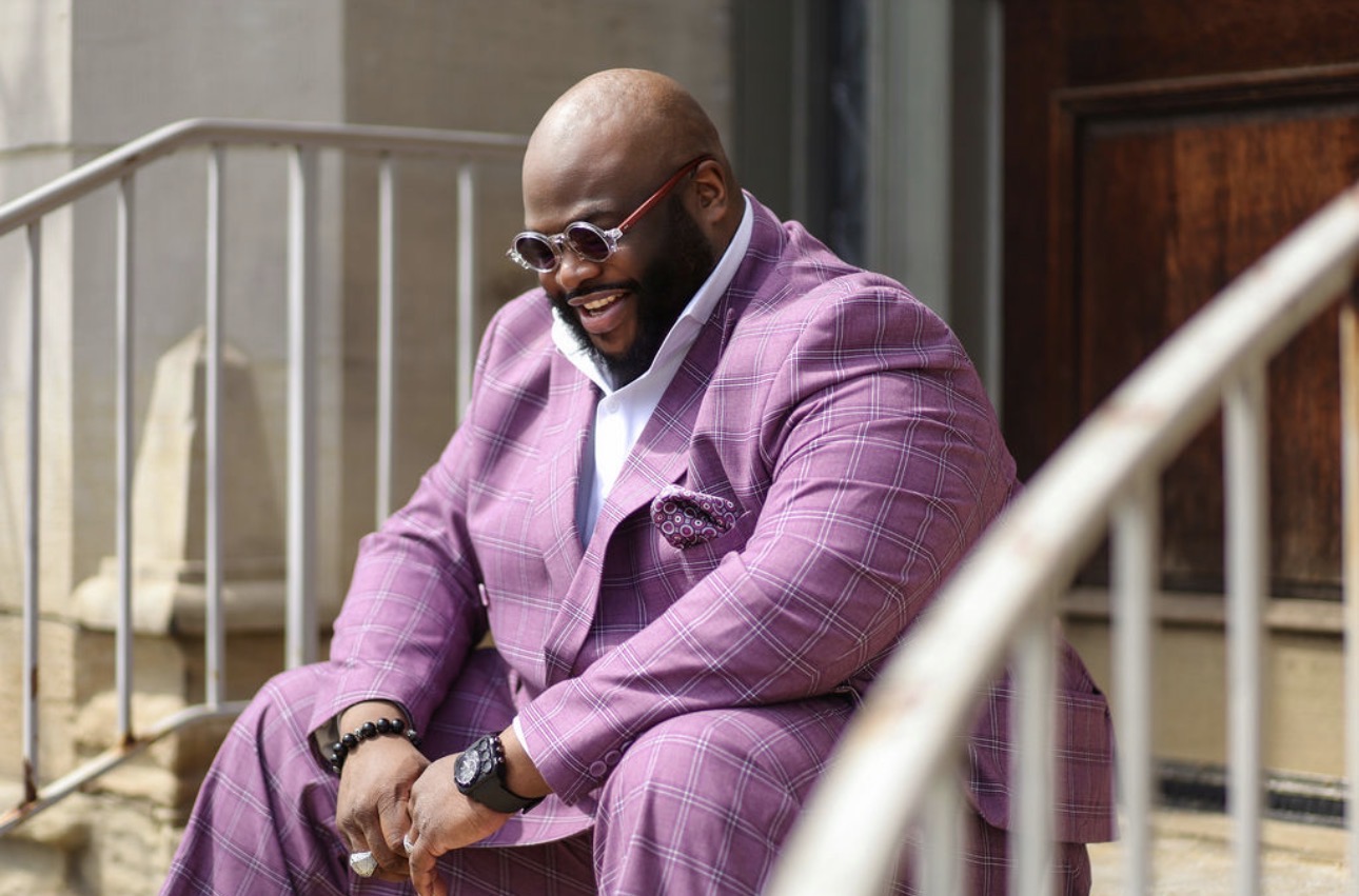 Man in purple suit sitting outdoors, smiling, wearing glasses and a watch.