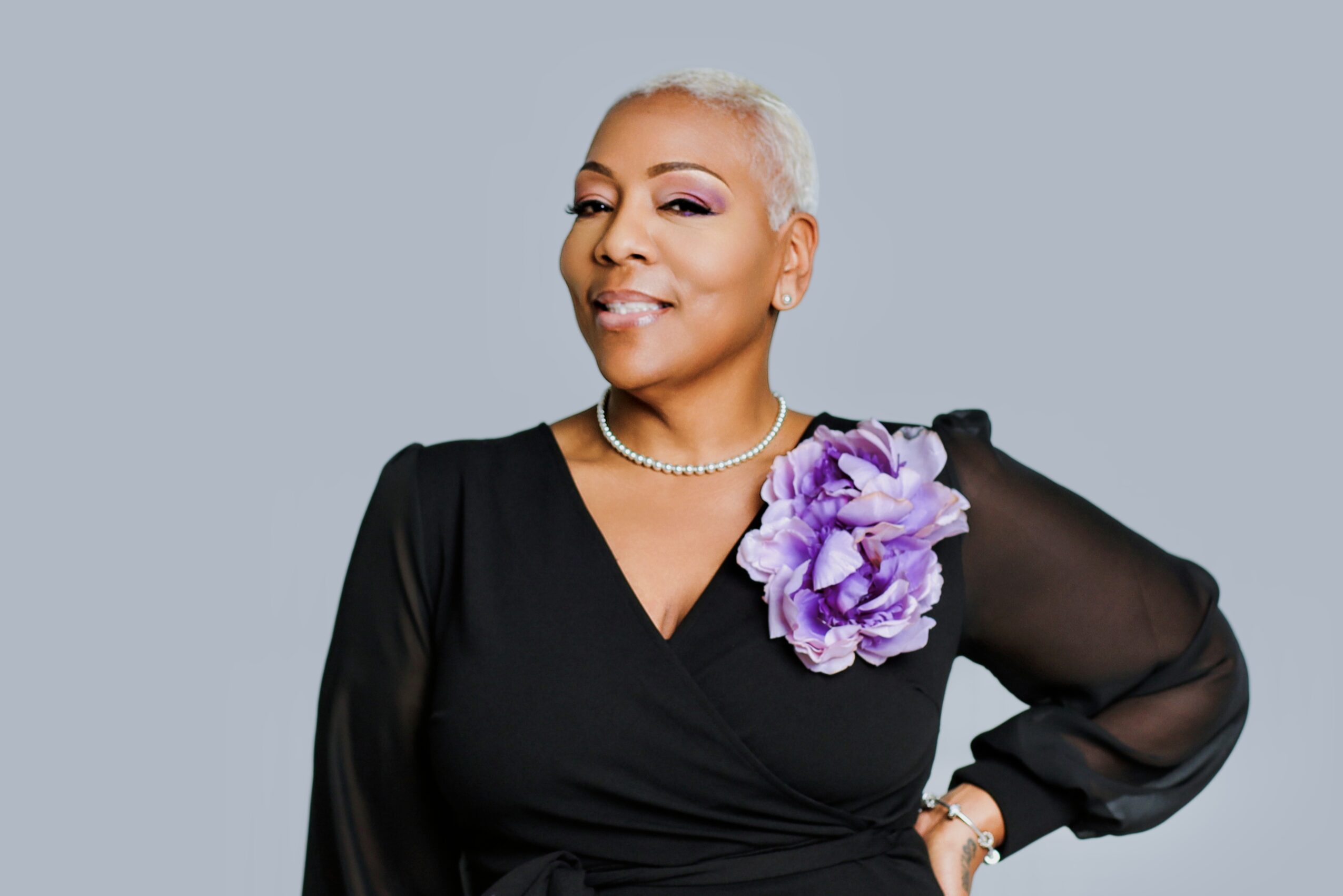 A woman with short gray hair wearing a black dress with a purple flower on her shoulder, smiling confidently.