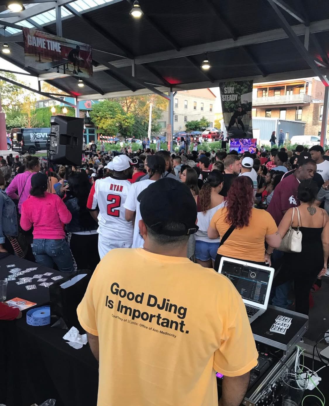 Crowd gathered at an outdoor event with a person in a yellow shirt in the foreground, facing away, with a laptop and sound equipment.