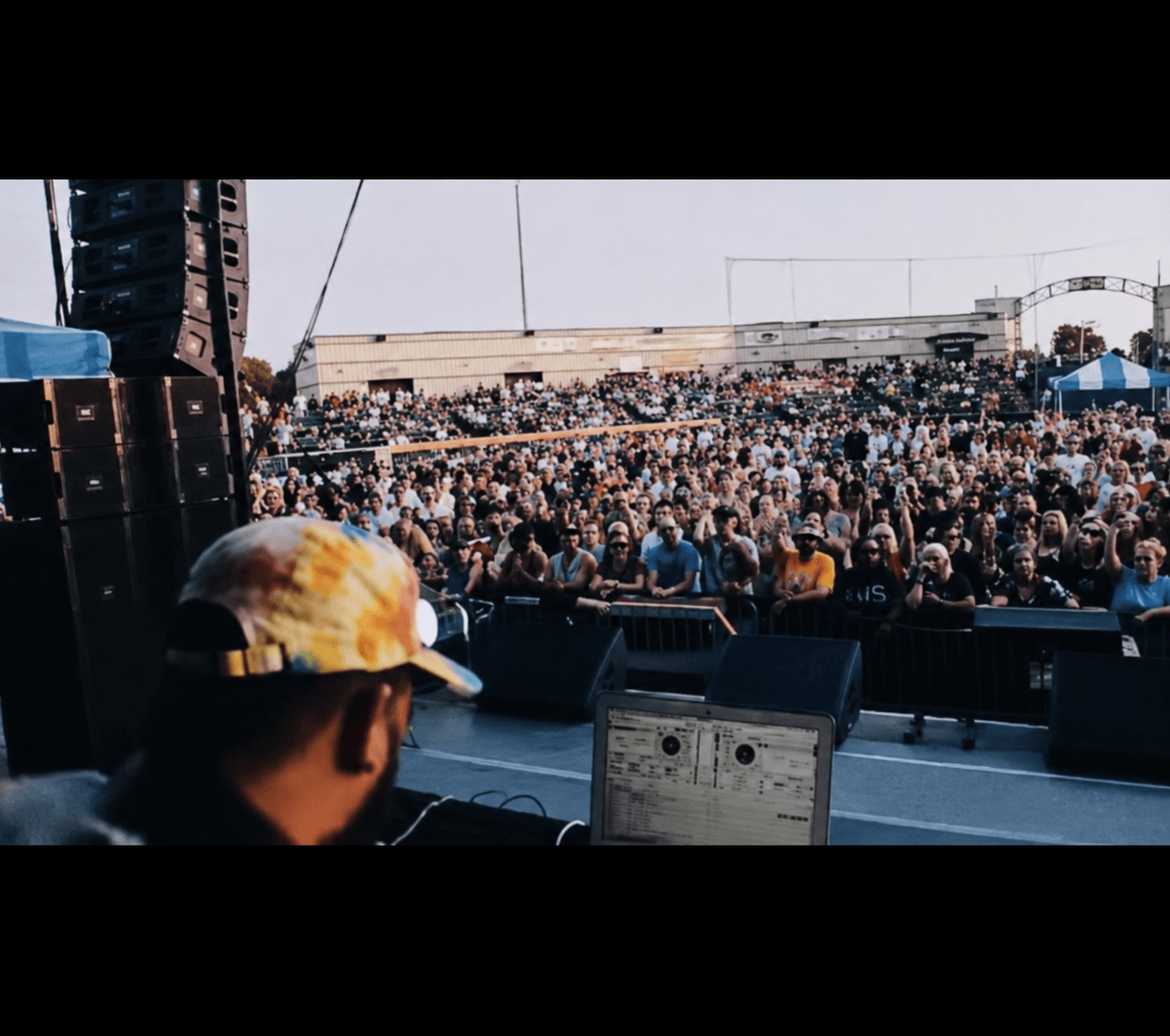 Crowd gathered outdoors at a concert with a DJ in the foreground, stage equipment, and a clear sky.