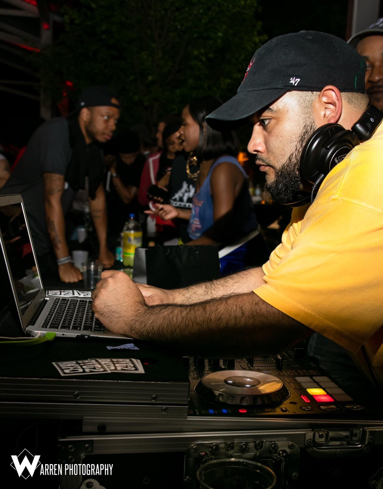 DJ wearing a black cap and yellow shirt operates equipment at an outdoor event with people in the background.