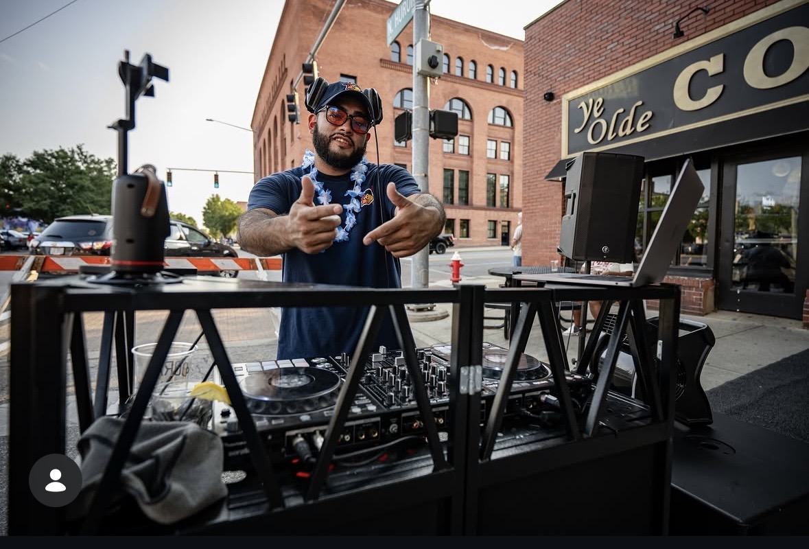 DJ with equipment outdoors in front of a building, smiling and pointing at the camera, with a city street background.
