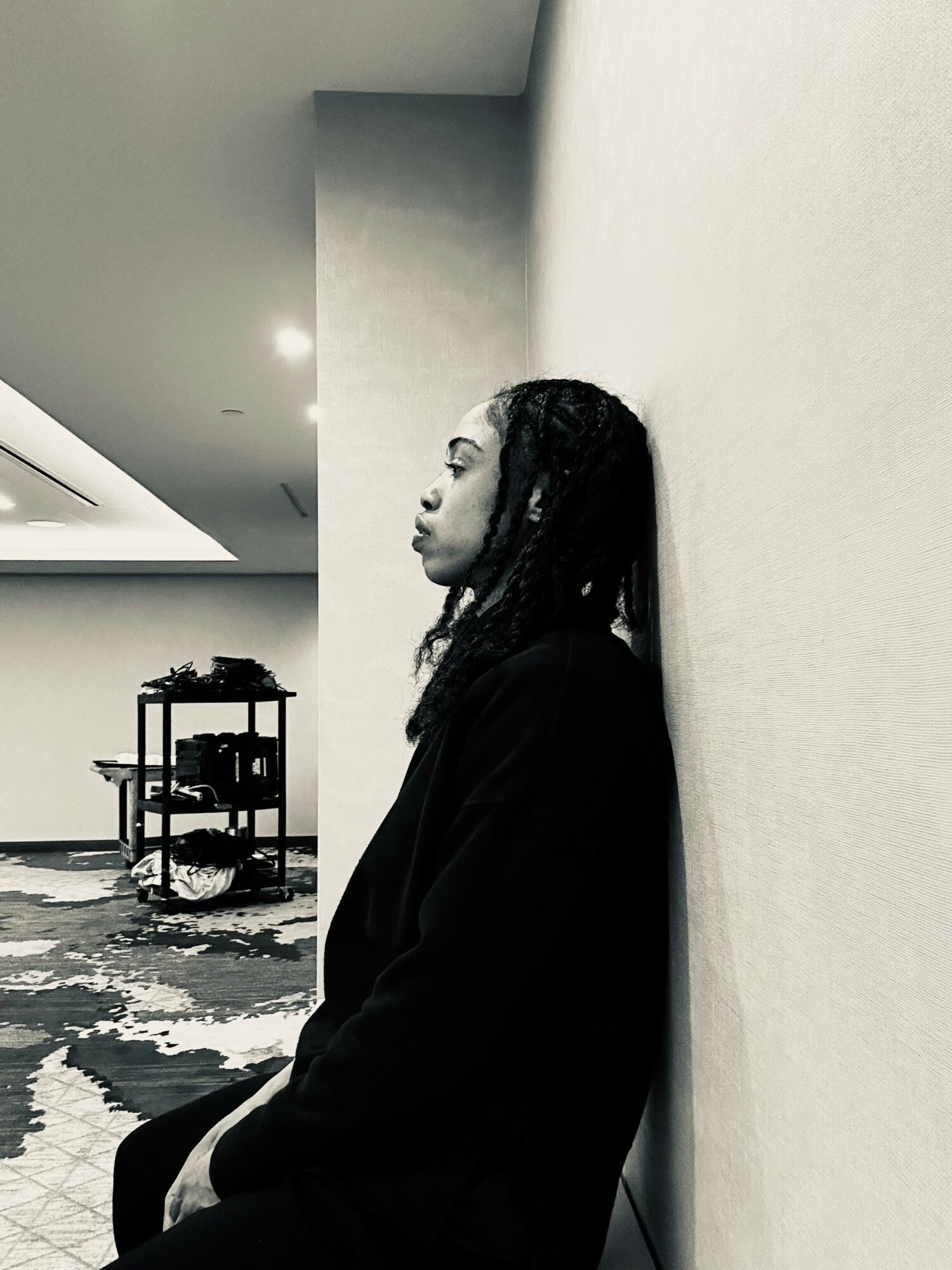 Person with long hair leaning against a wall in an indoor space, with a shelf and patterned carpet visible.