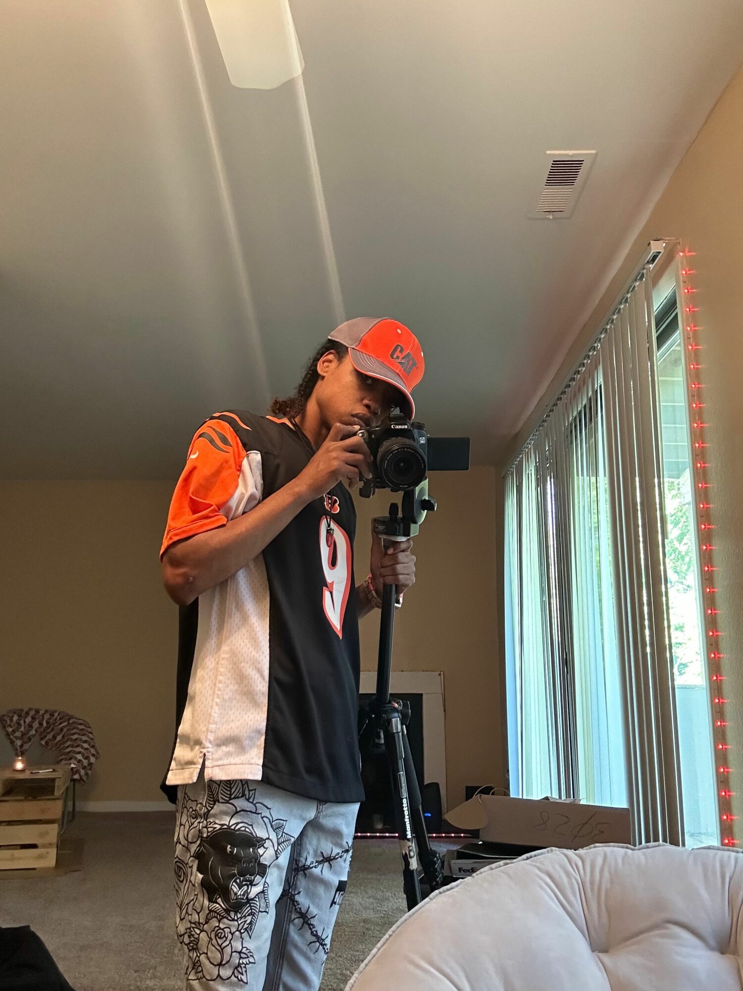 Person taking a mirror selfie in a room with a window and furniture, wearing a black and orange shirt and a cap.