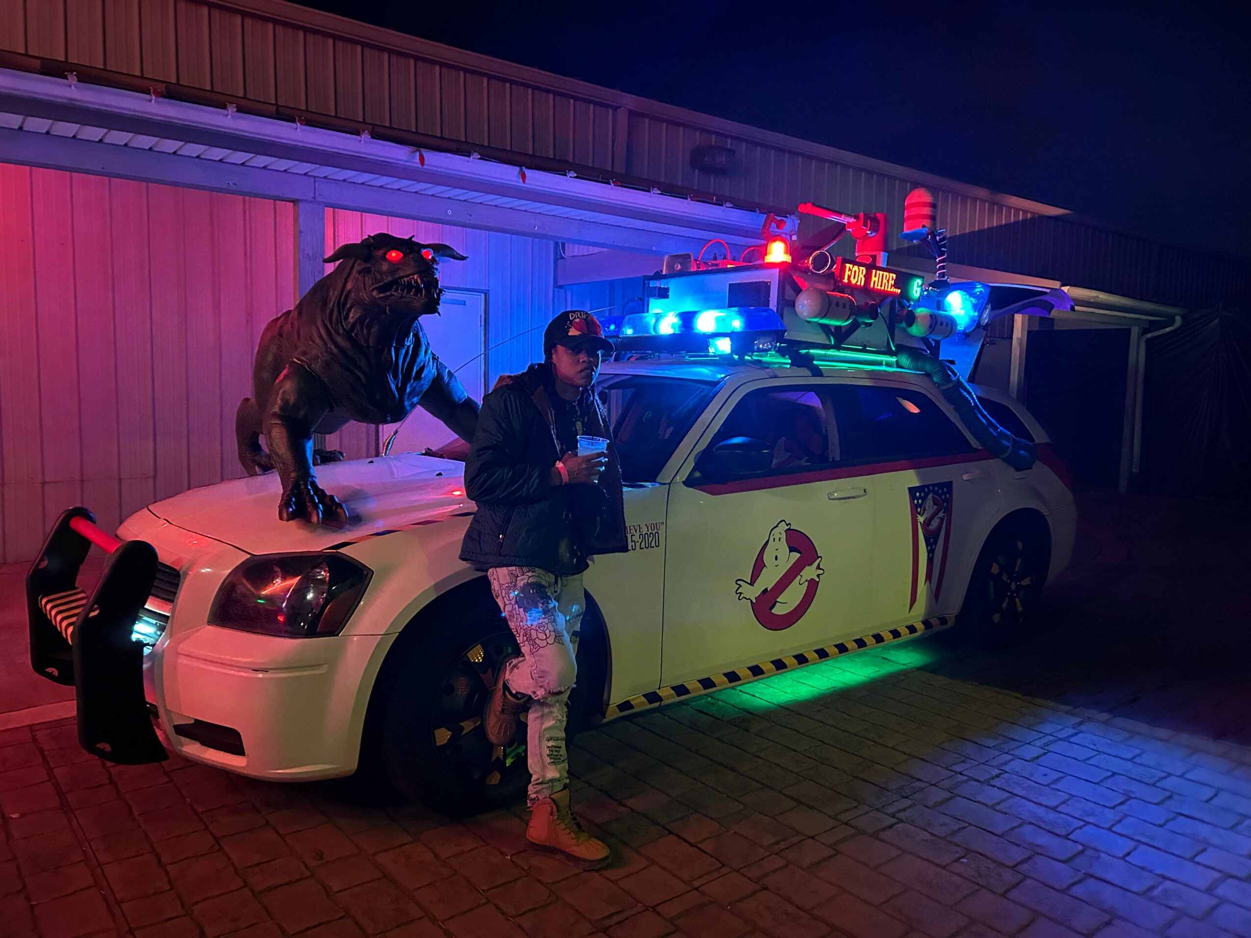 Two people and a dog pose in front of a police car with flashing lights at night, illuminated by colorful lights.
