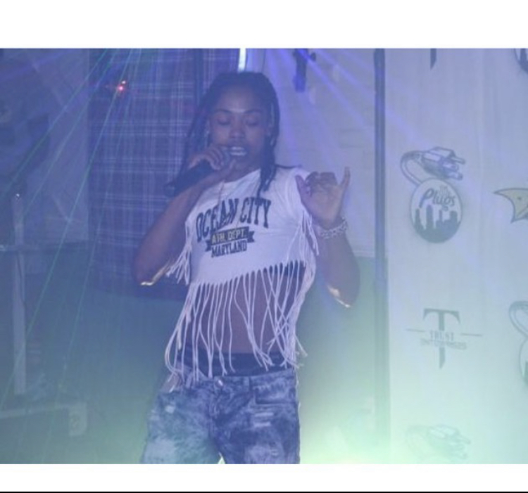 Person singing or speaking into a microphone on stage with a background of logos and lights.