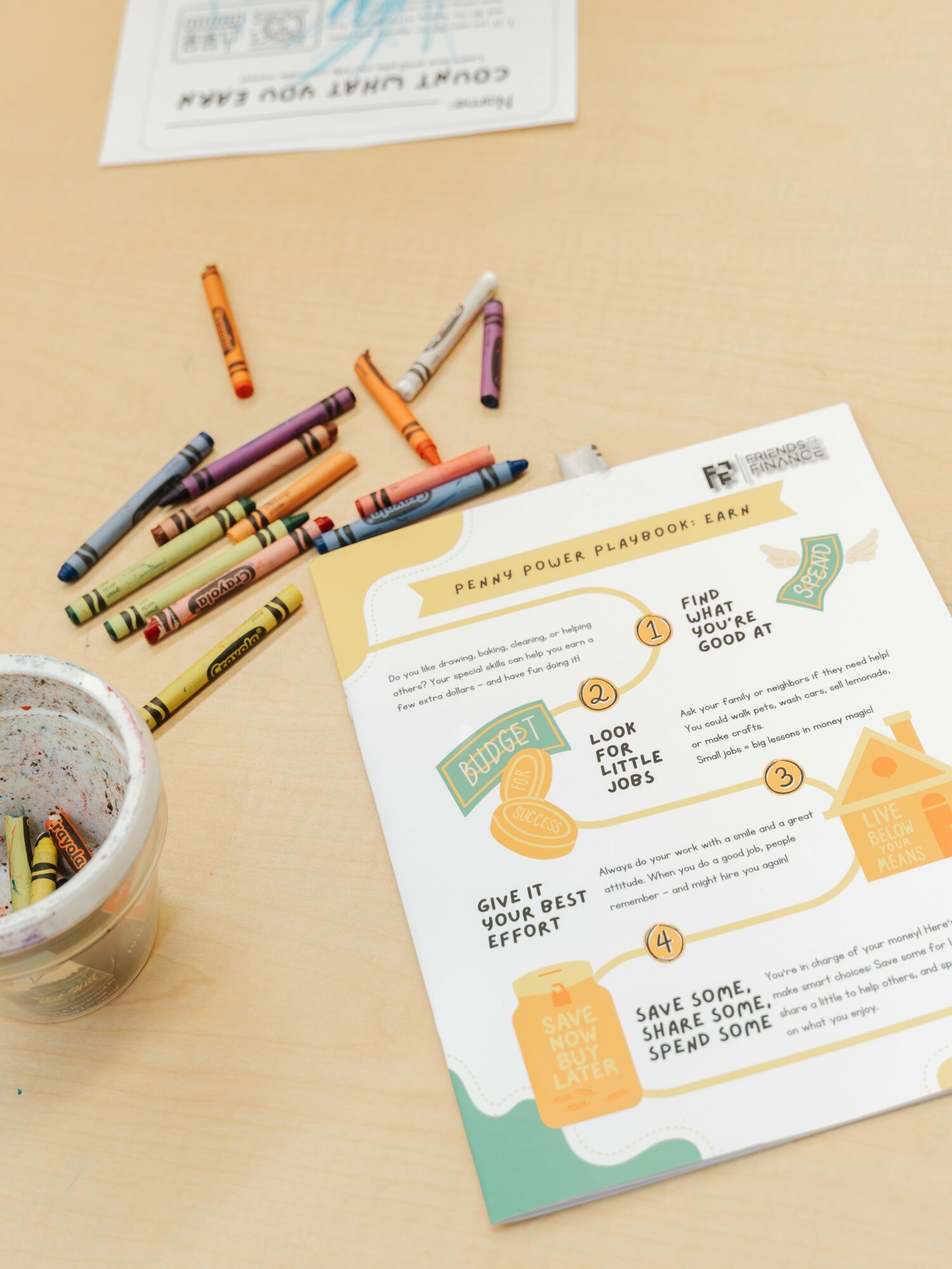 A worksheet with colorful illustrations and text, a container with pens, and a paper on a light wooden surface.