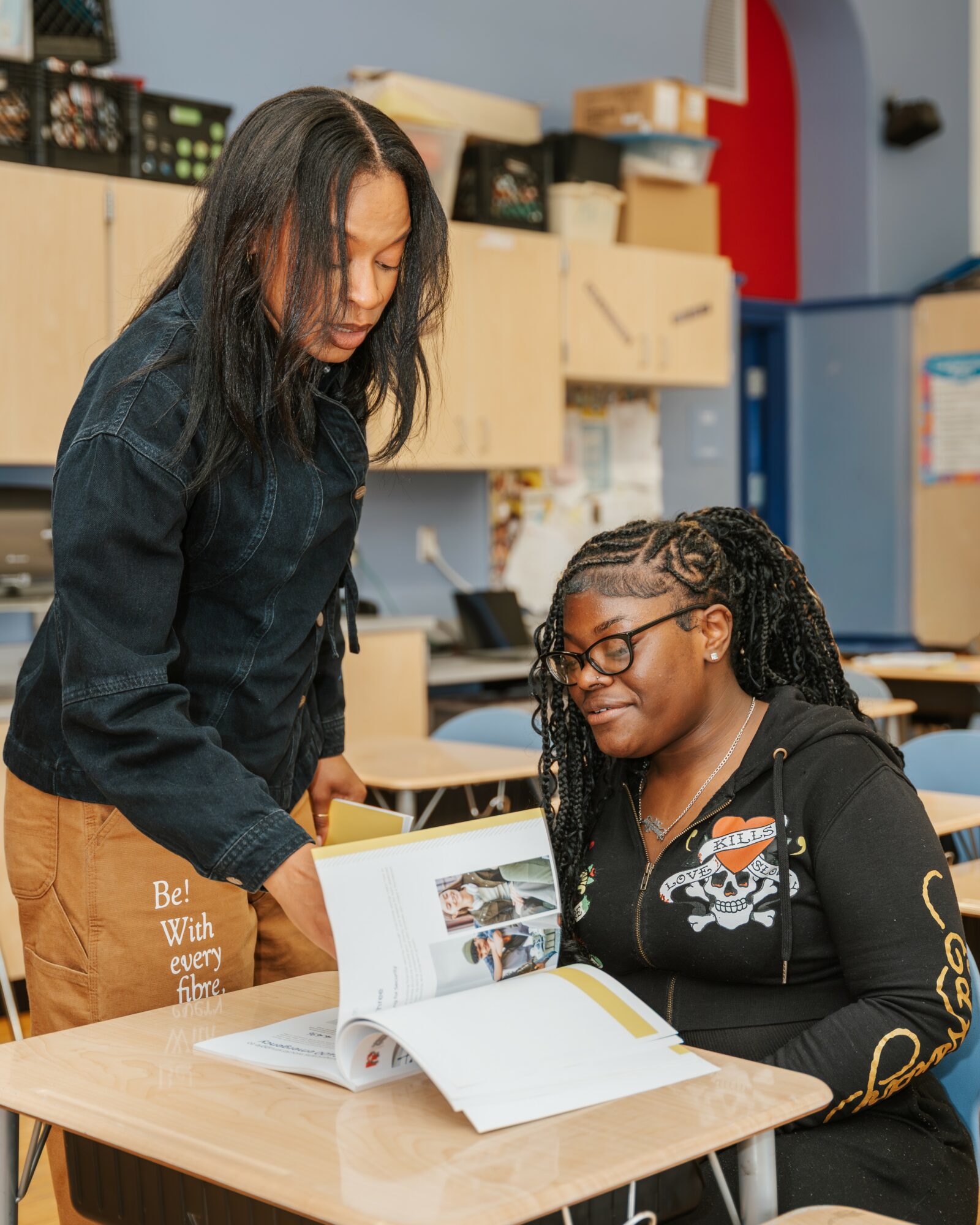 Two women look at a book on a desk in a classroom, with shelves and boxes in the background.