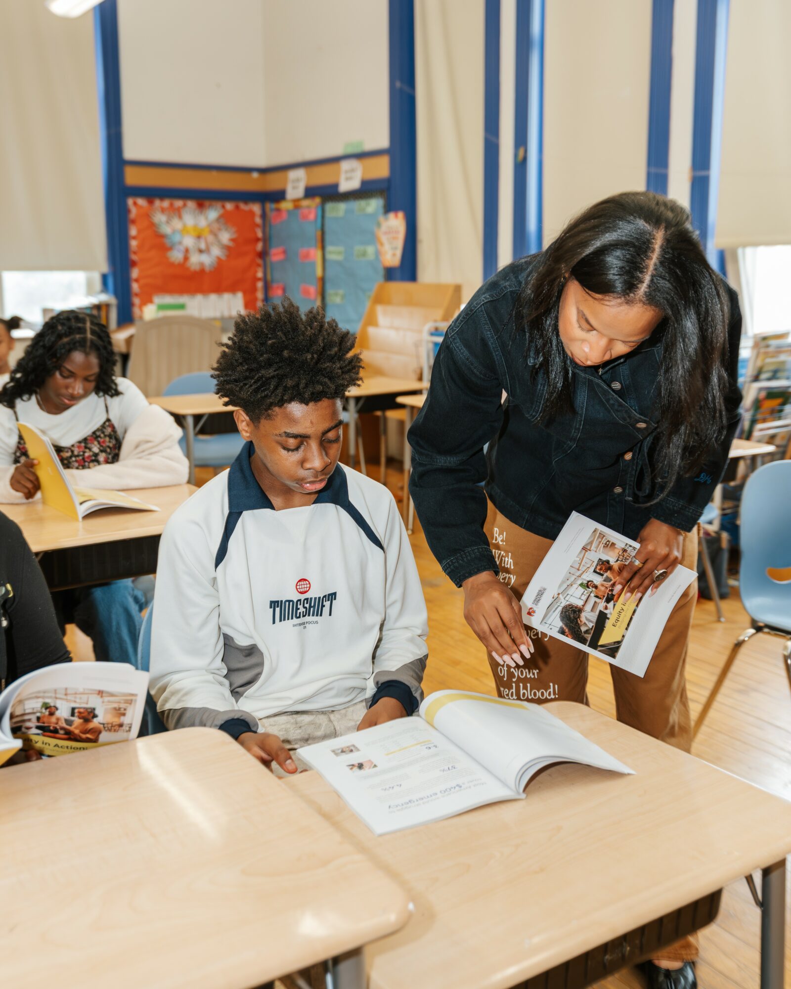 Teacher showing a book to a student in a classroom with other students, colorful decorations on the wall, and desks.