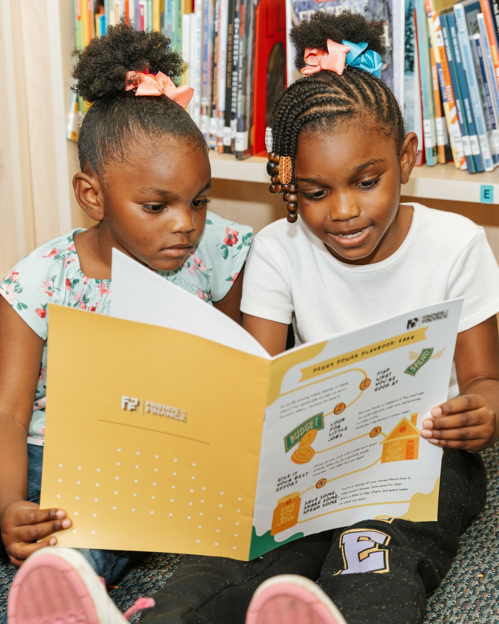 Two young girls sitting in a library, reading a colorful educational booklet together, with bookshelves in the background.