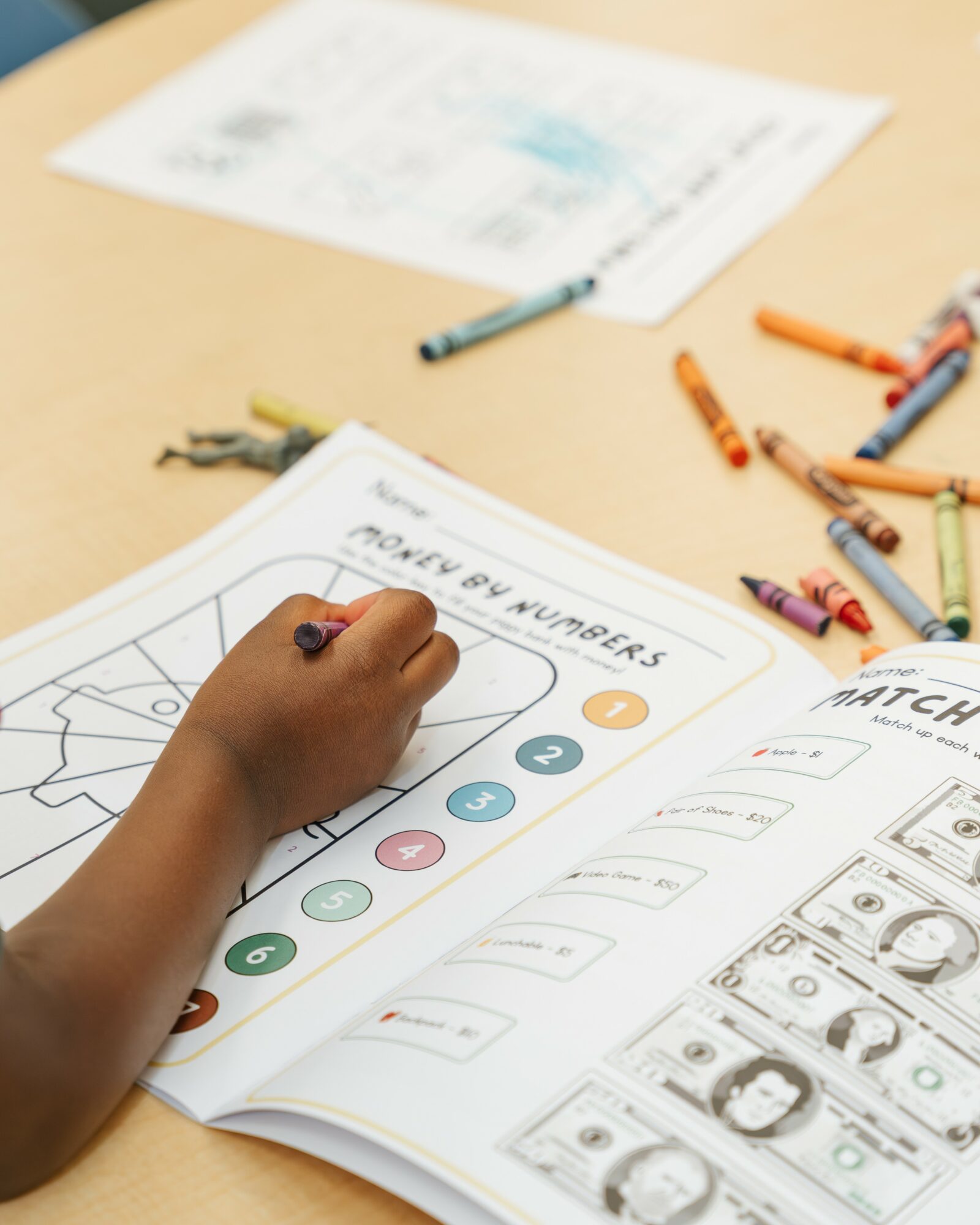 Child's hand coloring on a worksheet with numbers, surrounded by crayons and papers on a table.