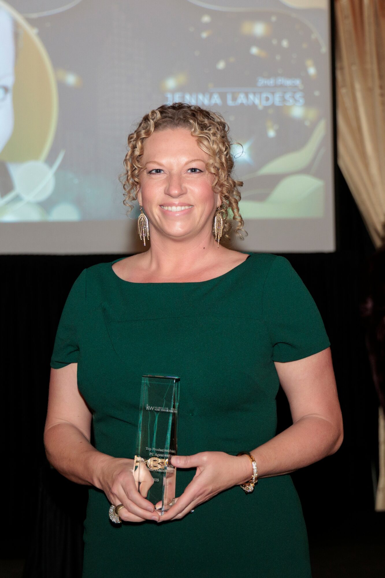 Woman with curly hair in a green dress holding a glass award, smiling, in front of a screen and curtain.