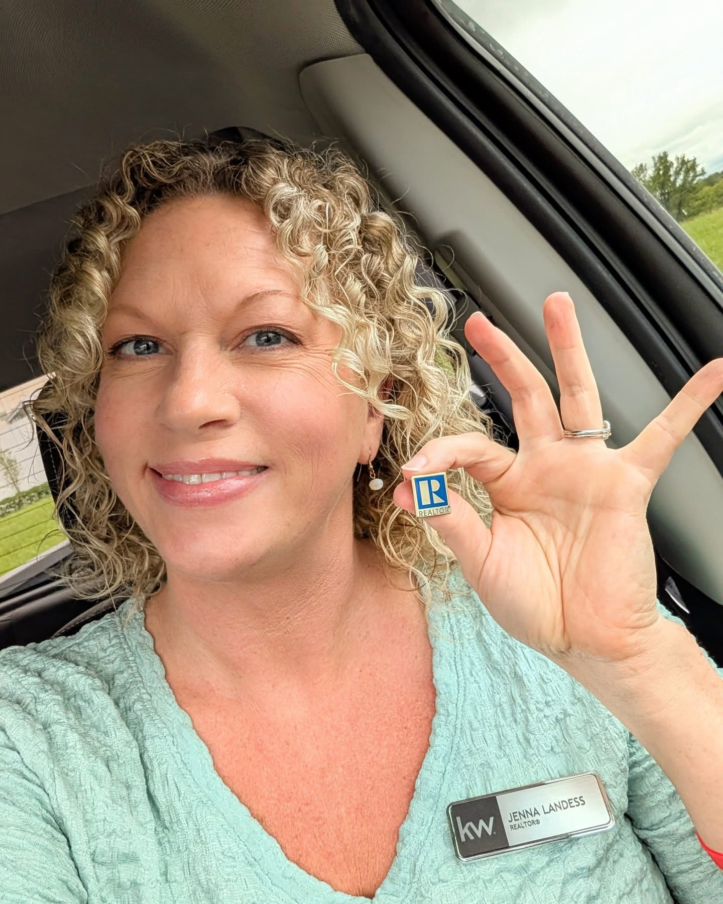 Woman with curly blonde hair smiling, holding a small blue and white object, inside a vehicle with a window showing greenery.