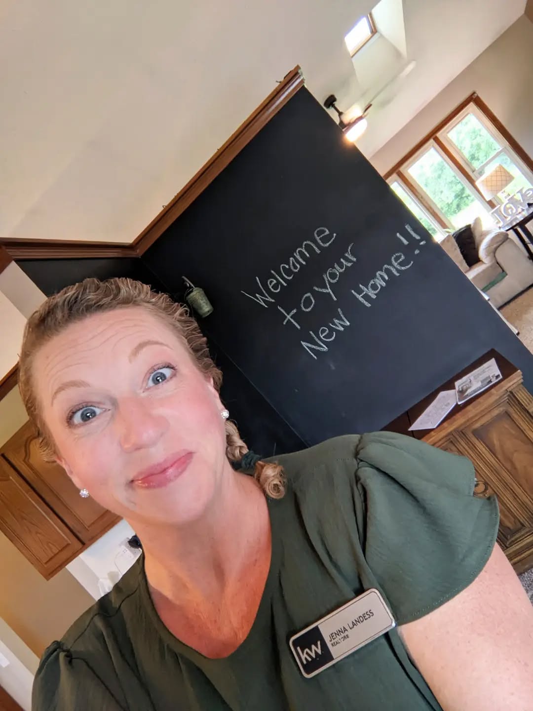 Woman with curly hair and green shirt smiling indoors, blackboard with welcoming message in background.