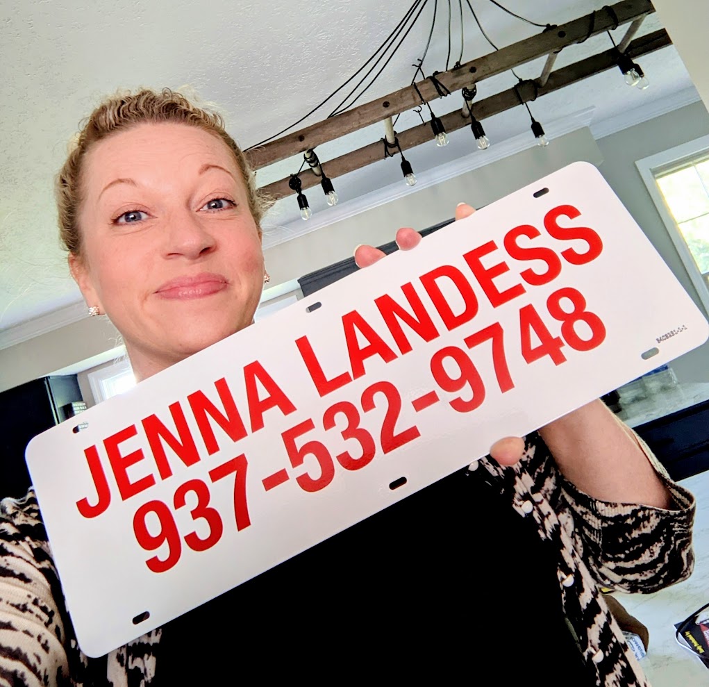 Woman holding a sign with her name and phone number, smiling, indoors with a window and ceiling lights visible.