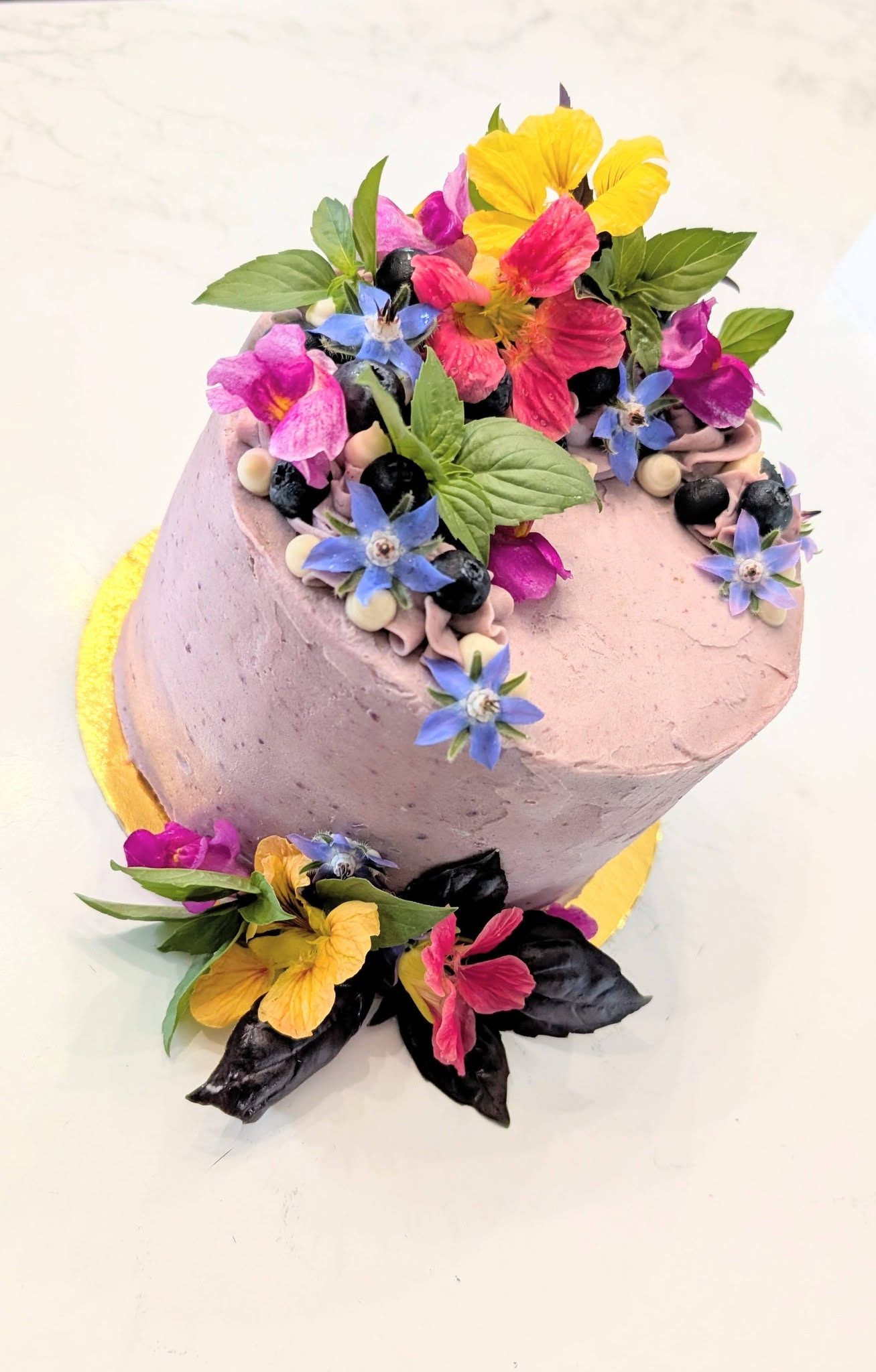 Round cake decorated with colorful flowers and green leaves, with a yellow base and black ribbon, on a white surface.