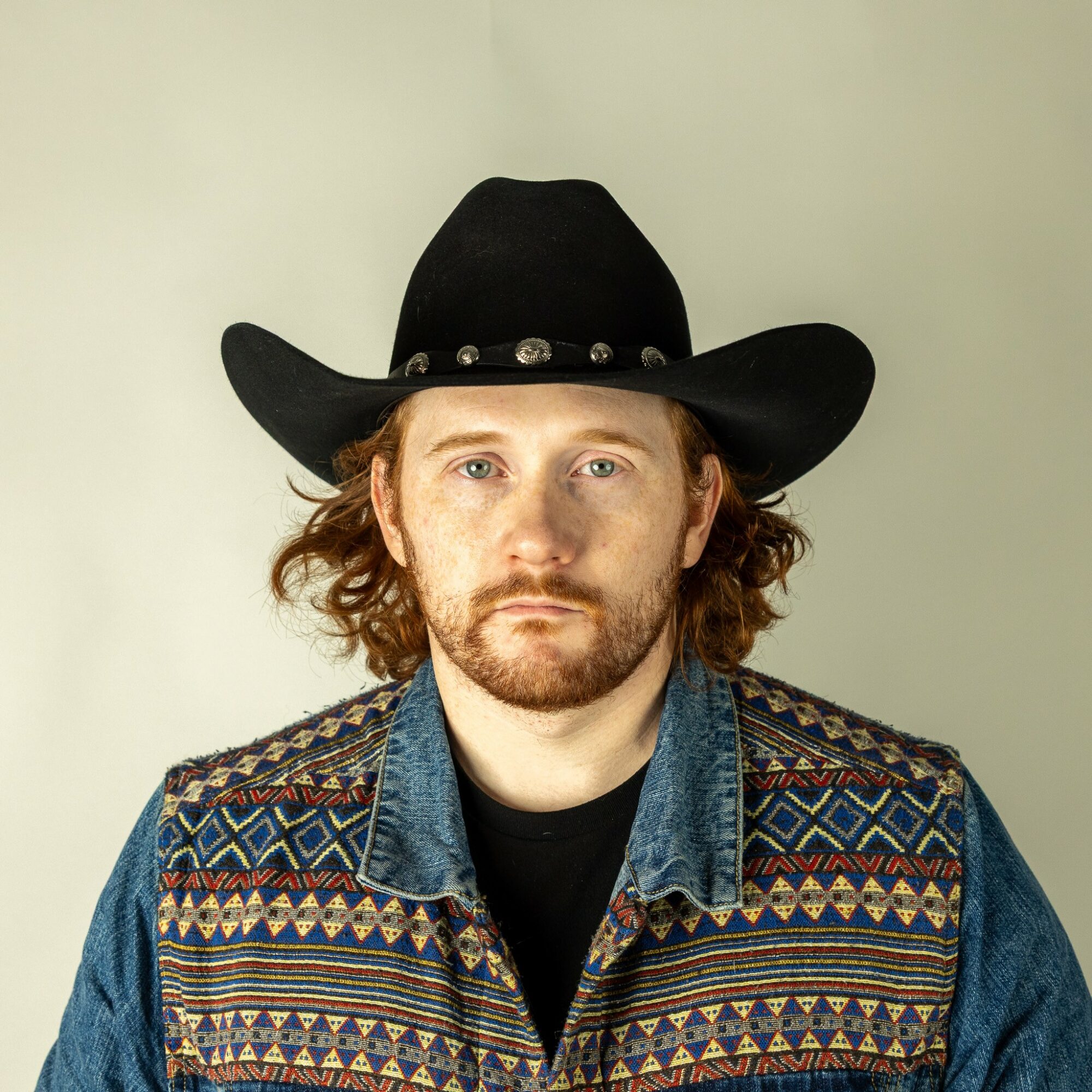 Man with curly hair and beard wearing a black cowboy hat and a colorful patterned jacket.
