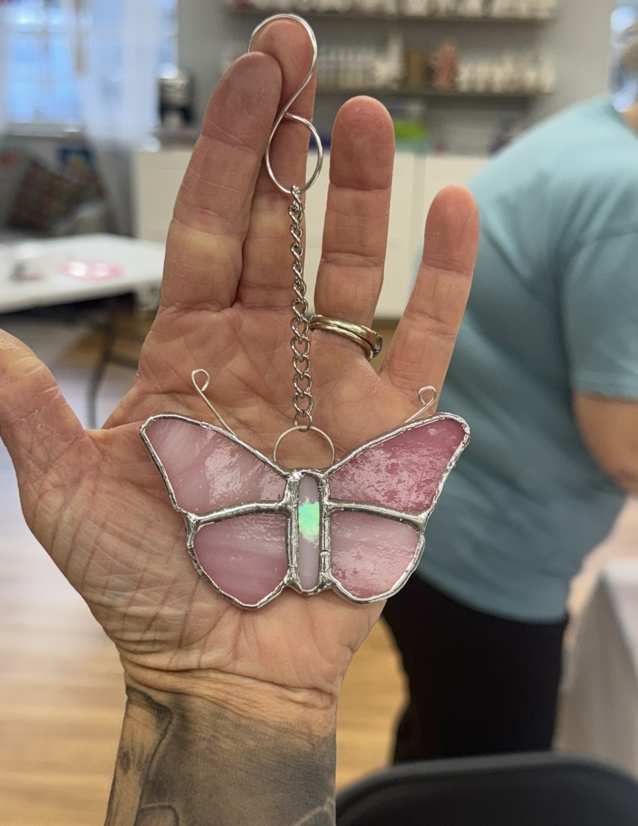Hand holding a butterfly-shaped pendant with pink wings and a green body, chain hanging from fingers, background of a room.