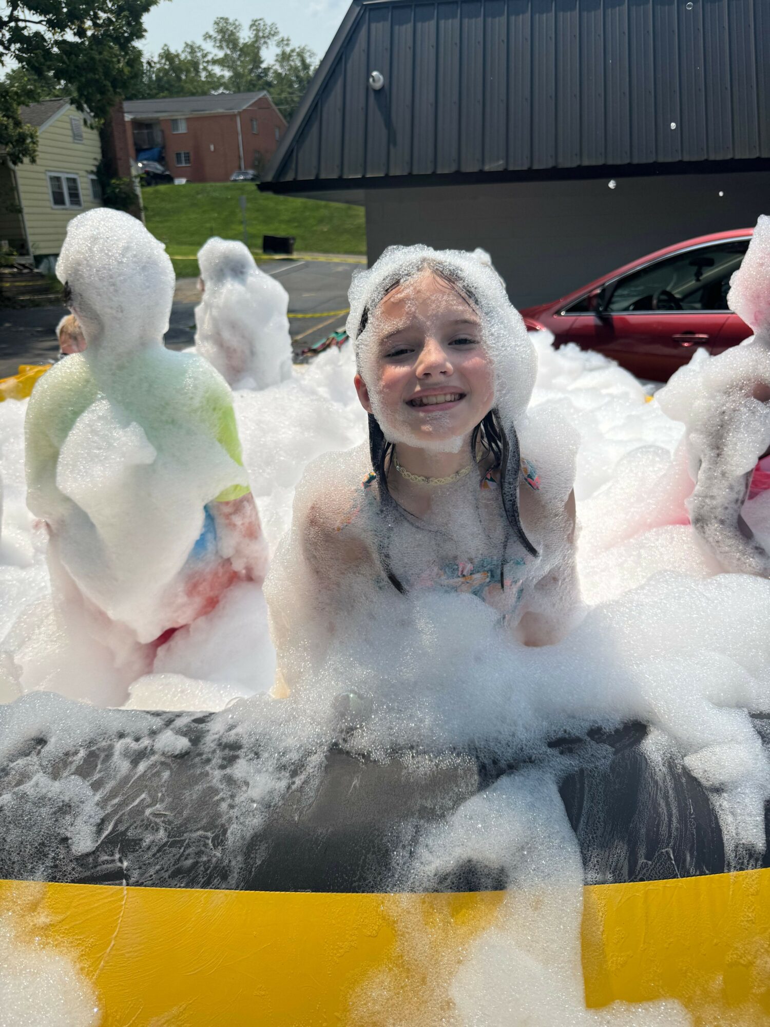 Person in a bathtub filled with bubbles, with a dog and a rubber duck, outdoors near a house and yard.