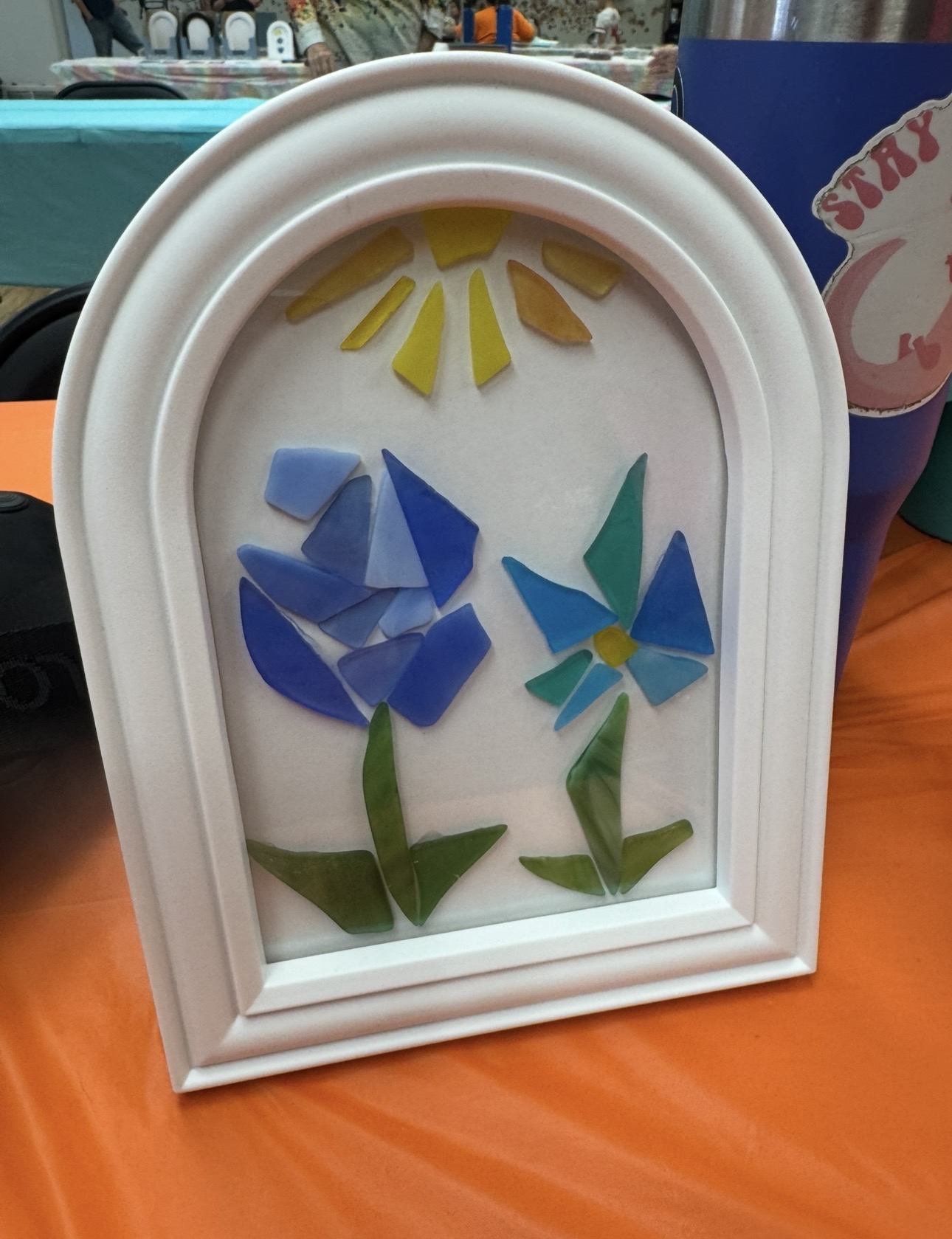 Decorative framed glass with colorful flower and sun mosaic design, placed on an orange surface.
