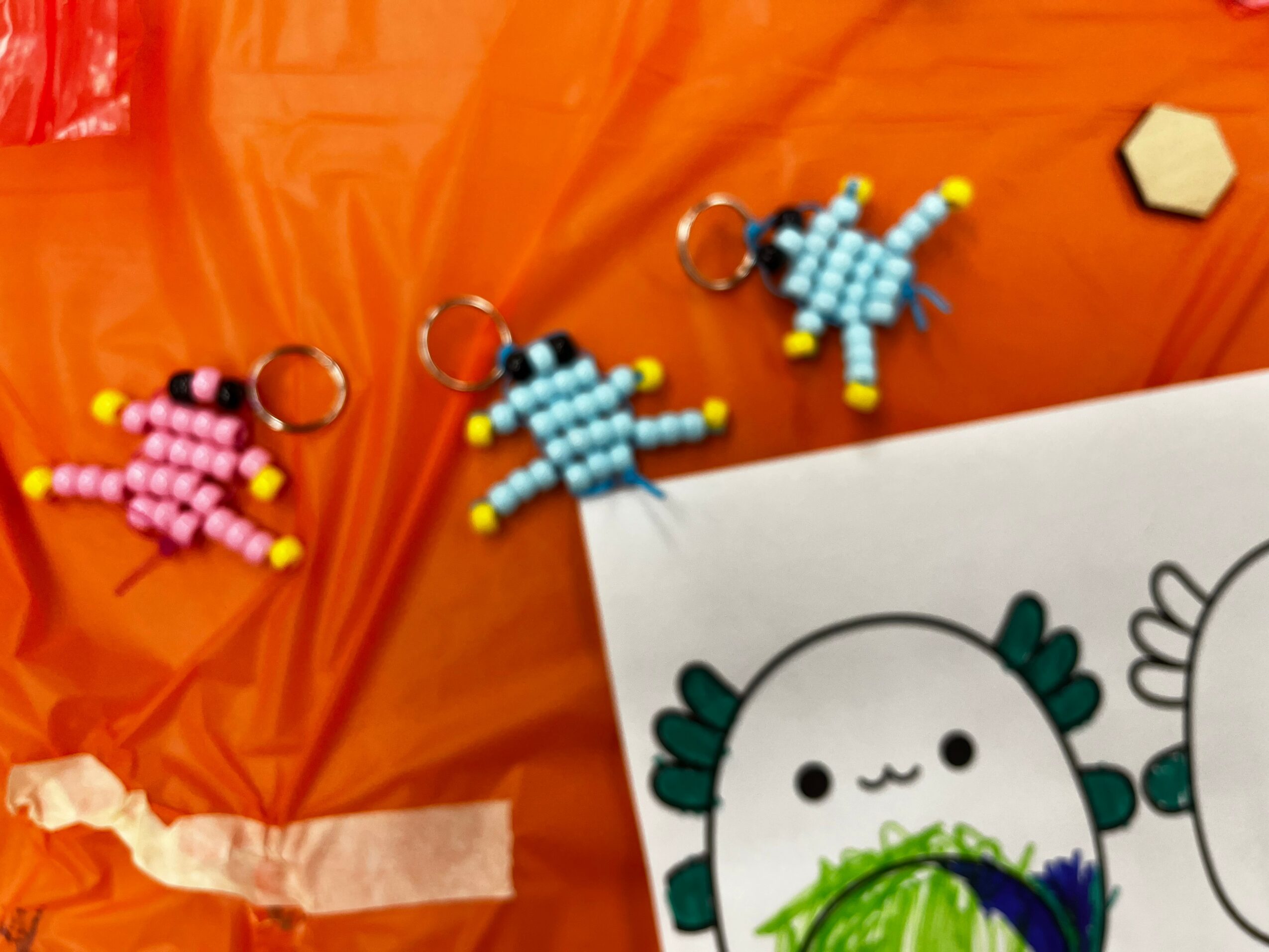 Three beaded keychains resembling animals on orange fabric, with a drawing of a character with a round face and green details.