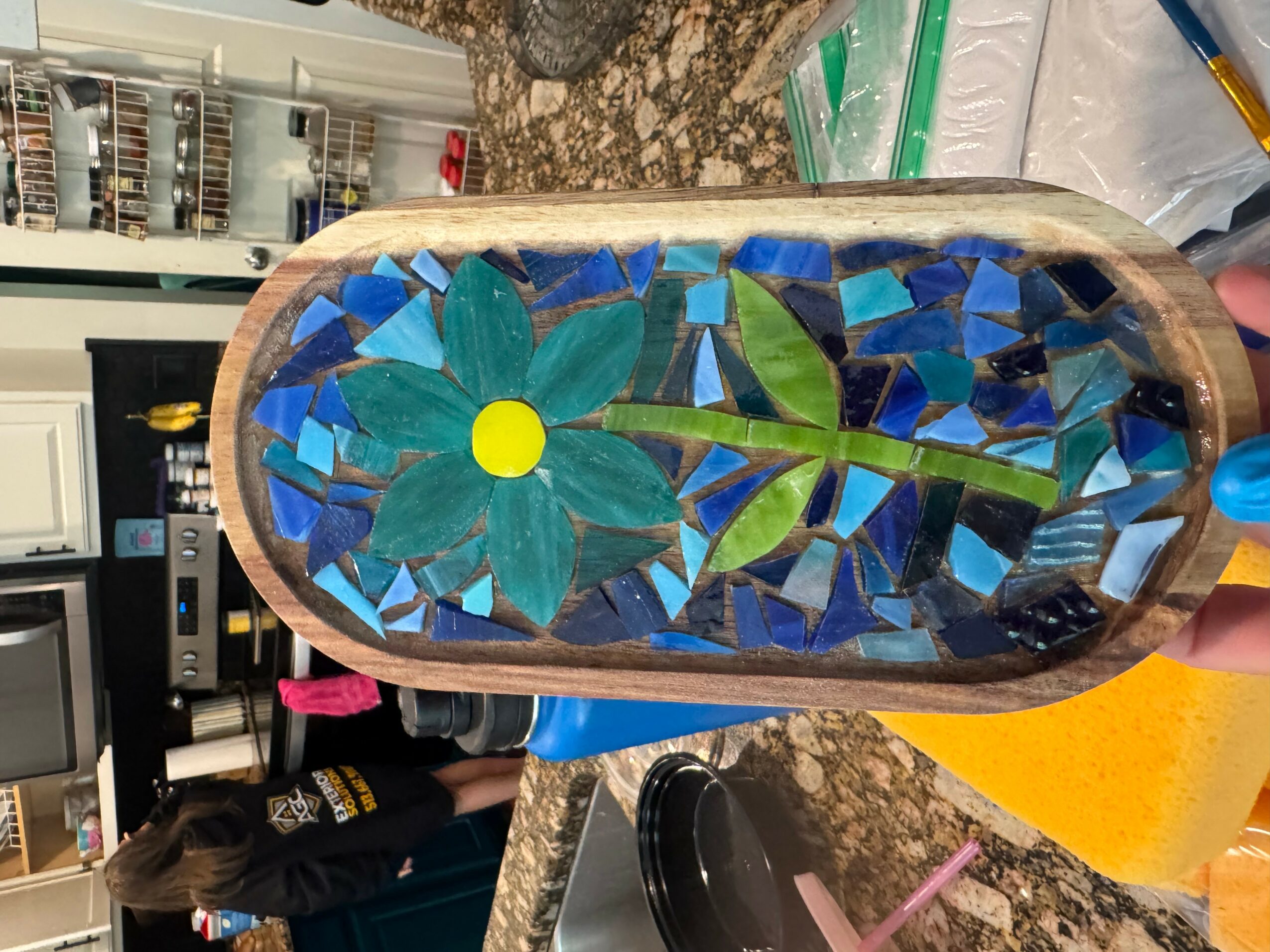 Colorful mosaic flower with green leaves inside an oval wooden frame, placed on a kitchen countertop.