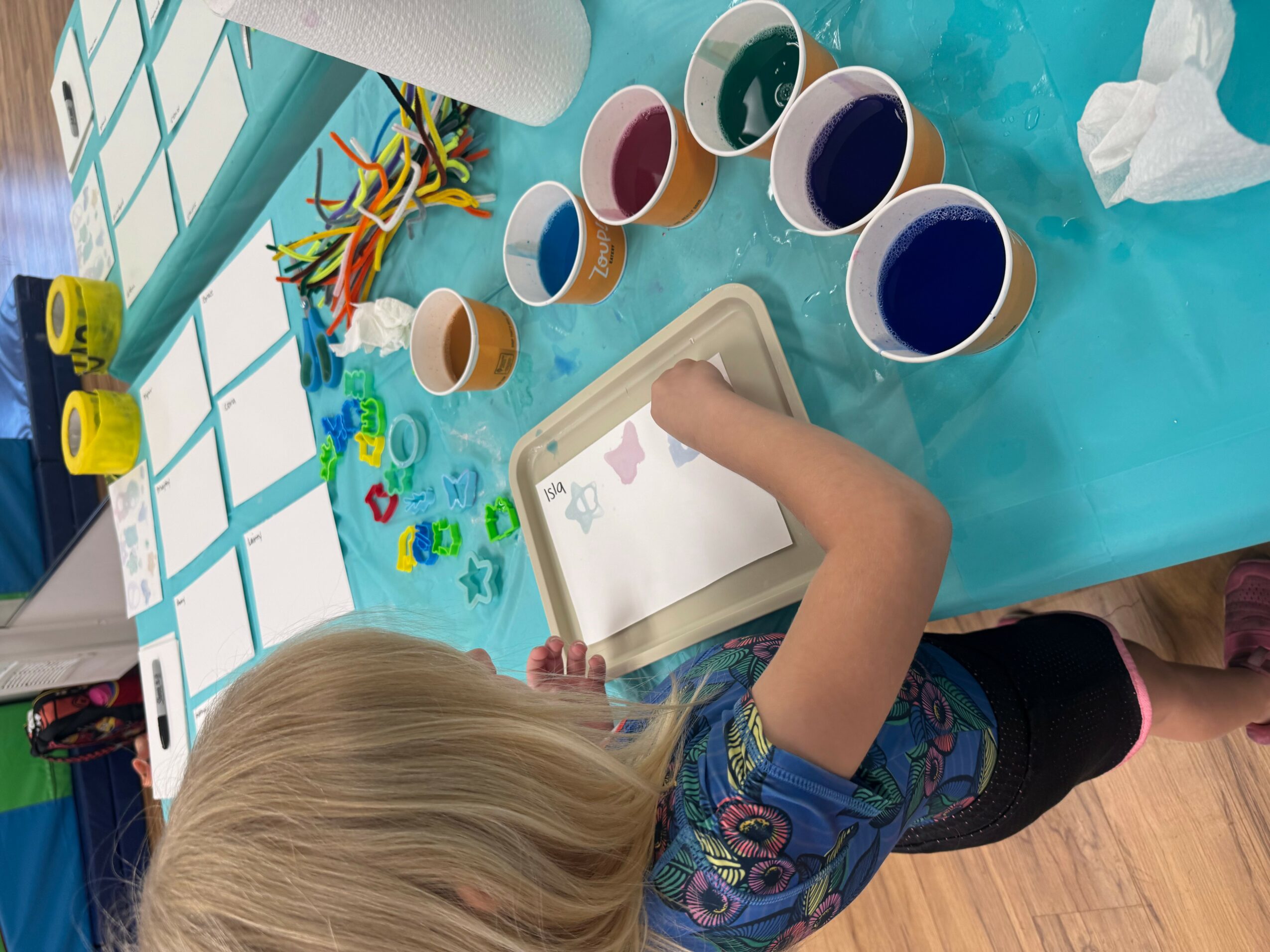 Child with blonde hair using a tablet at a table with cups of paint, craft supplies, and paper, in a bright room.