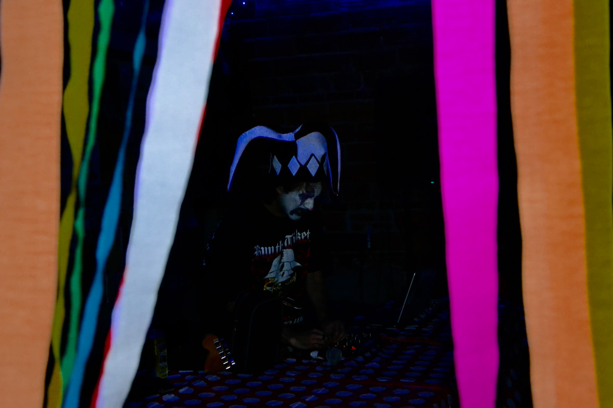 Dark figure with pigtails and diamond pattern on face, surrounded by colorful vertical stripes, in a dark setting.